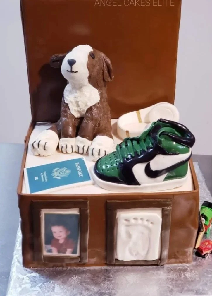 A birthday cake designed to look like a shoebox with a dog figurine, a pair of sneakers, a toy police car, a passport, and a framed photo on the sides.