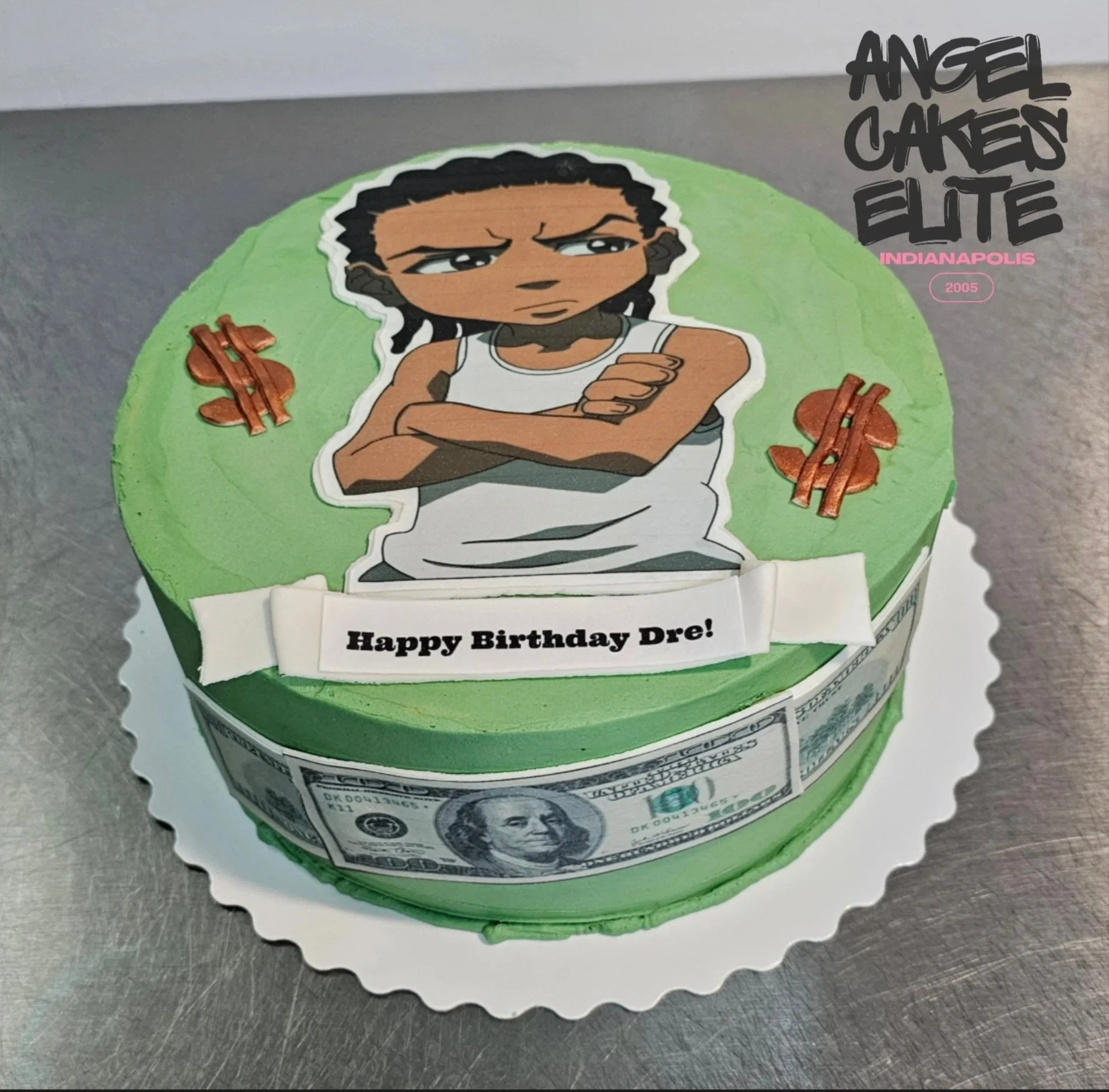 Birthday cake with green frosting, featuring a cartoon with braided hair, crossing h arms with an annoyed expression, surrounded by copper-colored dollar sign decorations. The cake has a white banner with black text saying, "Happy Birthday Dre!" and a band of printed dollar bill images around its side.