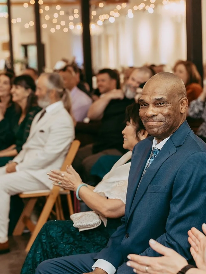 A diverse group of people dressed in formal attire sitting in an audience at an event, with warm lighting and string lights overhead.