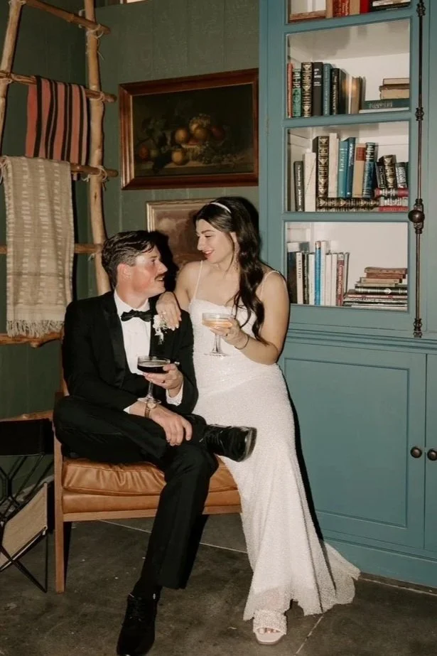 Bride and Groom in library holding glasses of champagne next to blue cabinet sitting on leather chairat the westport in downtown chico