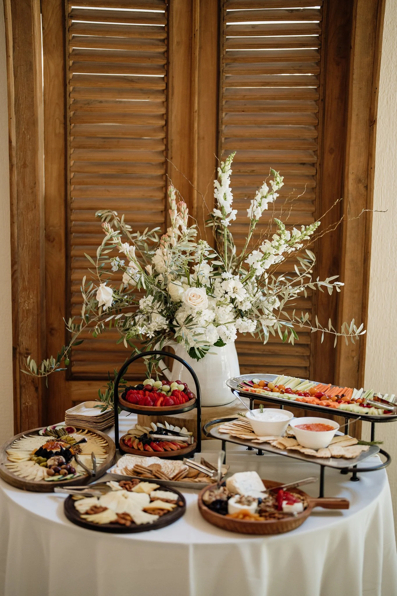 Buffet table with cheese, crackers, fruit, and dips, decorated with a large white floral arrangement on a white tablecloth, set against wooden shutters.