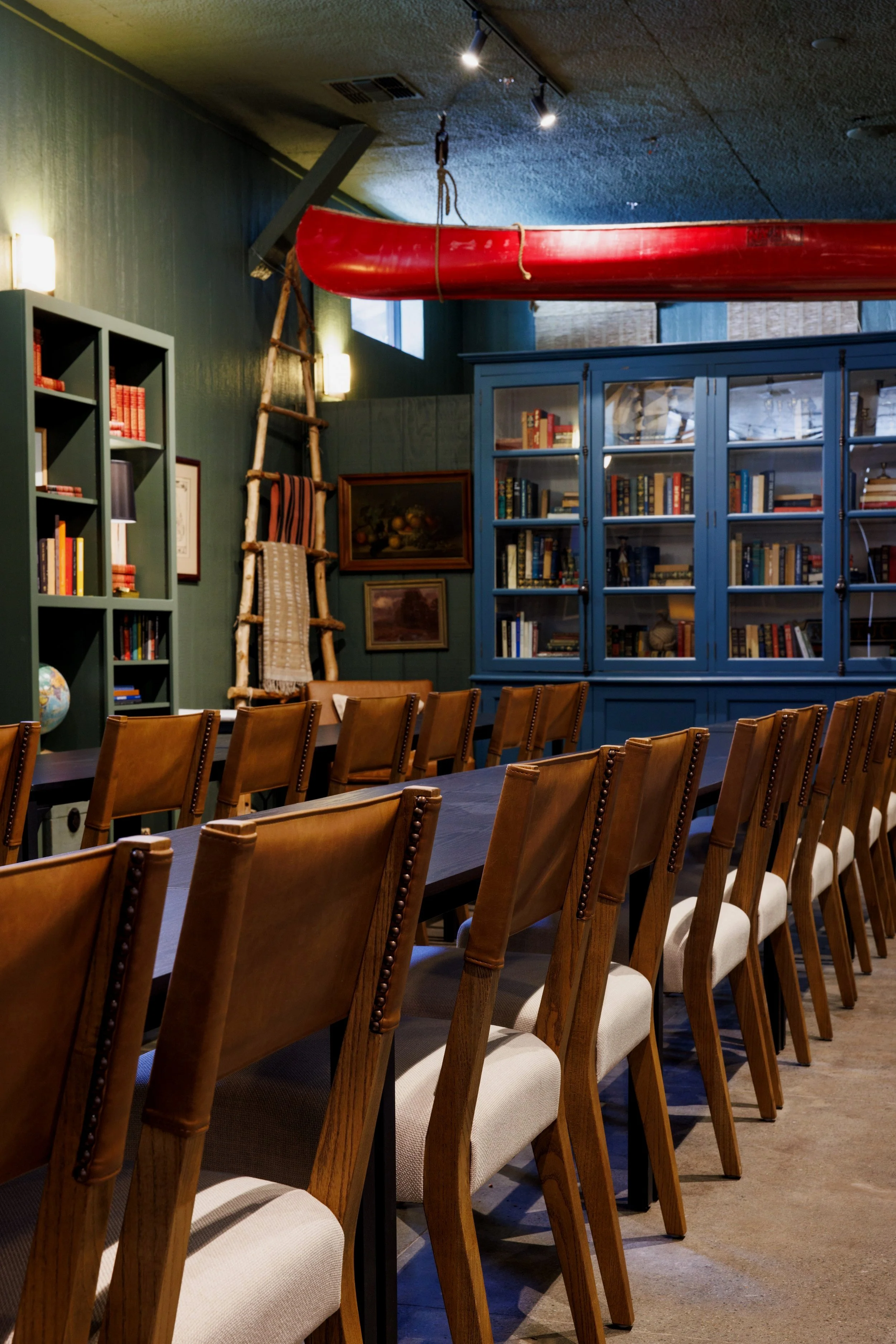 A meeting or dining room with a long wooden table and multiple brown upholstered chairs, green walls, a large blue bookshelf with glass doors filled with books, a red canoe hung from the ceiling, a ladder decorated with blankets, and paintings on the wall.