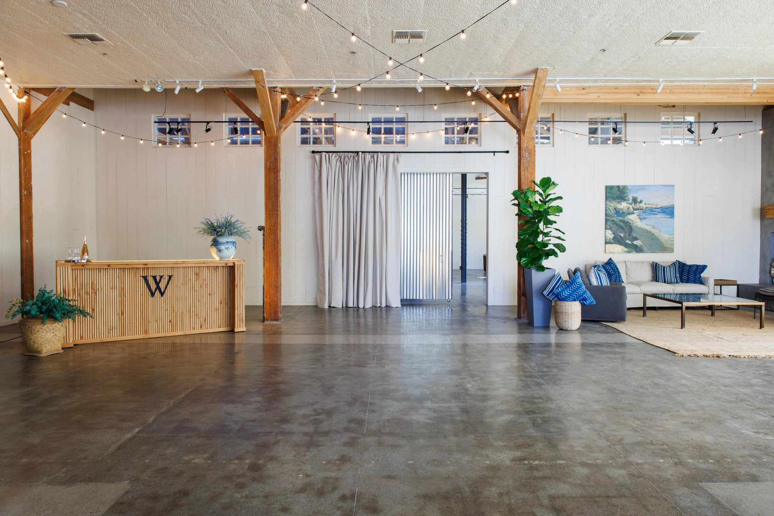 Interior of a modern event space with polished concrete floor, white walls, wooden beams, string lights, a reception desk with a large letter W, a sitting area with a beige couch and blue accent pillows, large potted plants, and artwork on the wall.