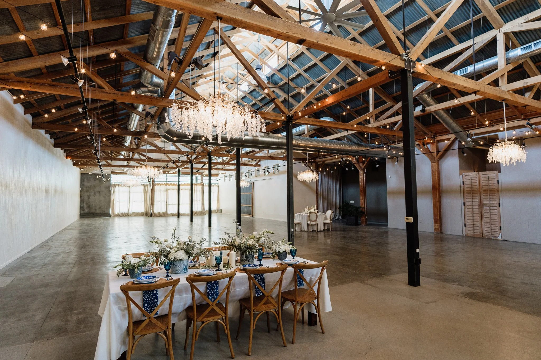 Decorated event hall with a long table set for a meal, with flowers and blue glasses, in a rustic space with wooden beams, chandeliers, and additional tables in the background.