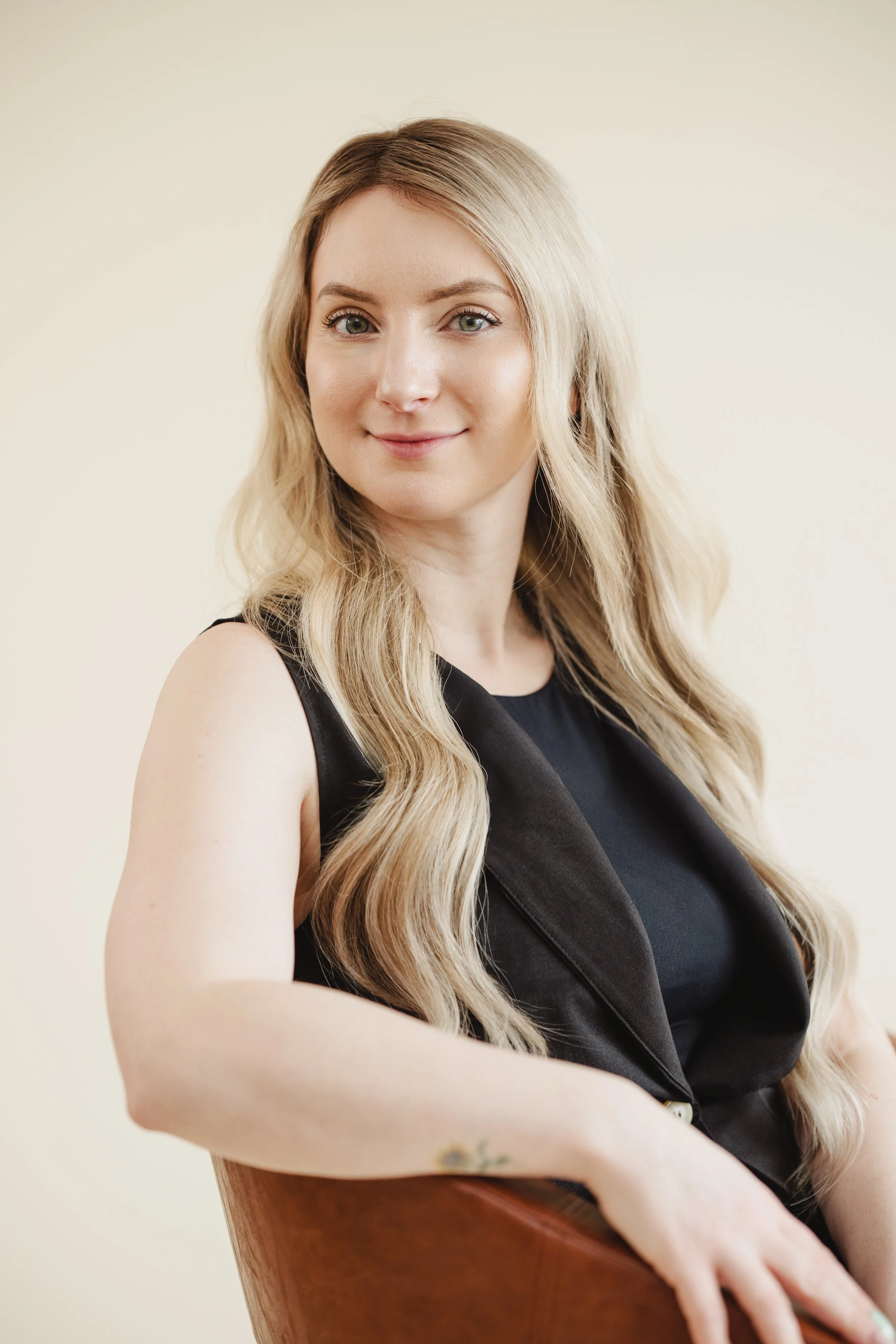 A portrait of a young woman with long, wavy blonde hair, wearing a black turtleneck sweater, standing outdoors in a narrow alleyway with brick and stucco walls.