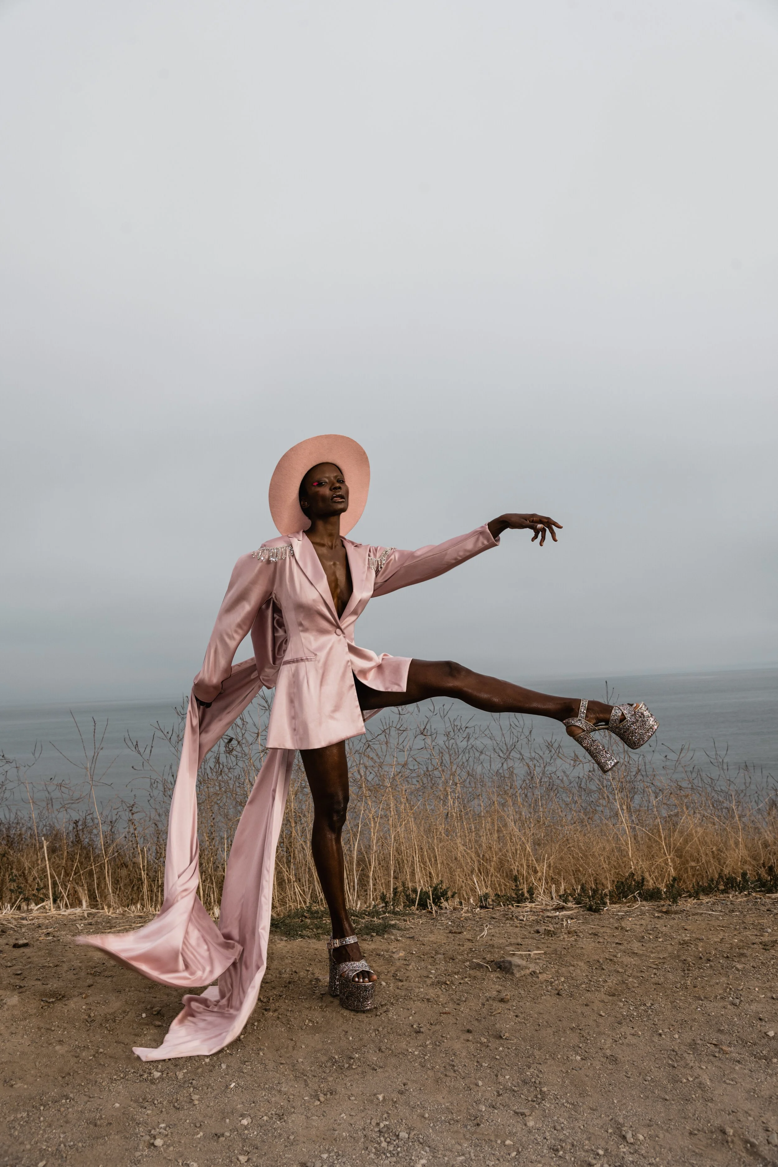 Person posing outdoors by the ocean, wearing a pink outfit with long sleeves, a wide-brim hat, and platform heels, standing on one leg with arms extended.