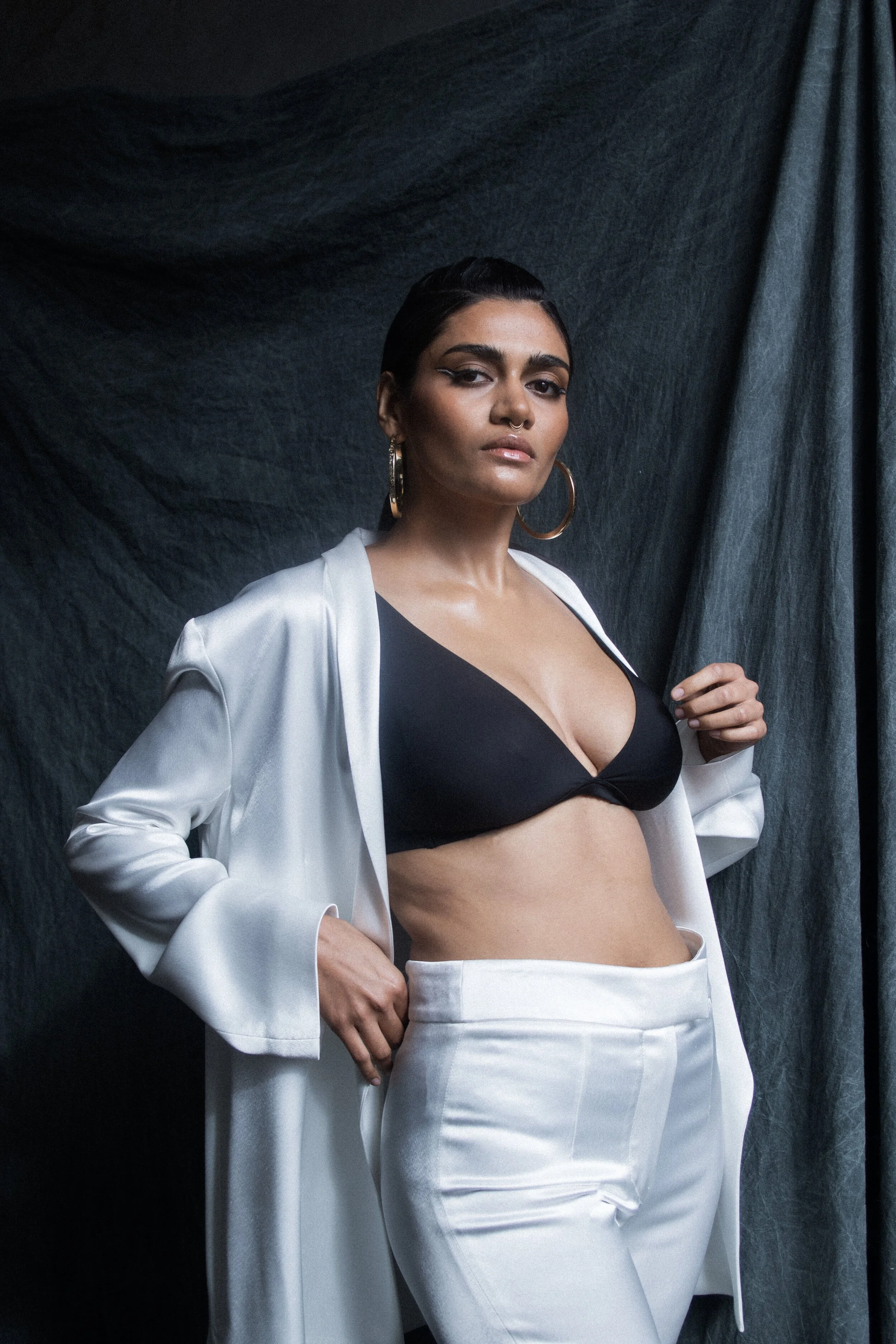 Person wearing a black bra and white suit with gold hoop earrings against a dark fabric backdrop.