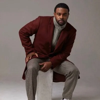 Man in a burgundy coat sitting on a stool against a gray background