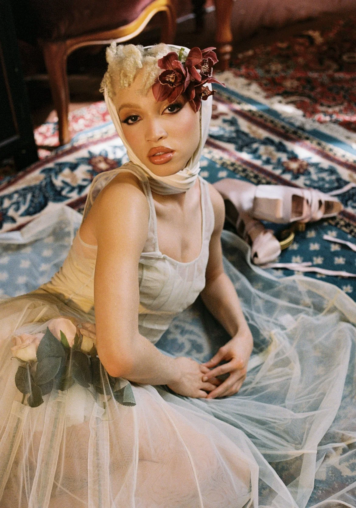 A person in an elegant dress sits on a patterned carpet. They wear a headscarf adorned with flowers. Nearby, there are ribbons and shoes. The scene is softly lit, creating a vintage, artistic ambiance.