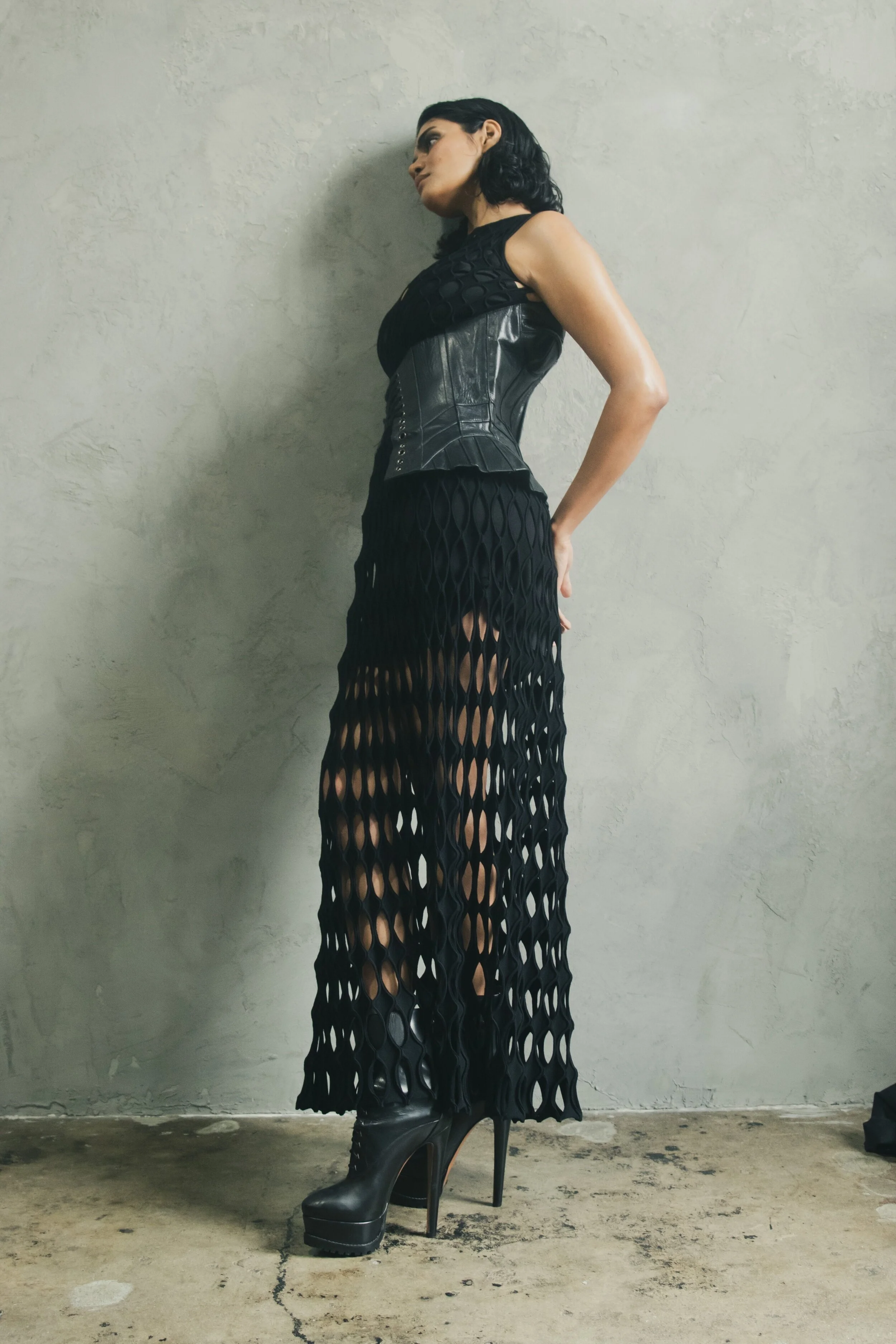 Person wearing a black leather corset, long mesh skirt, and platform boots, leaning against a textured wall.