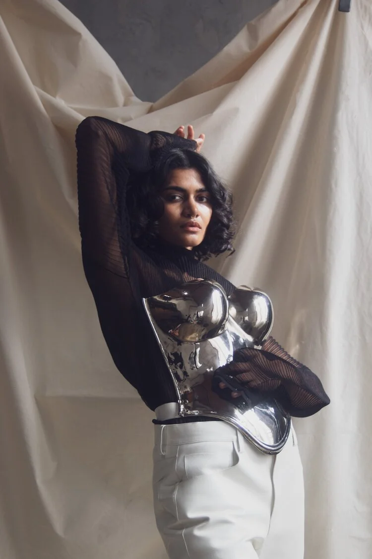 Person posing in a metallic corset with sheer black top and white pants.