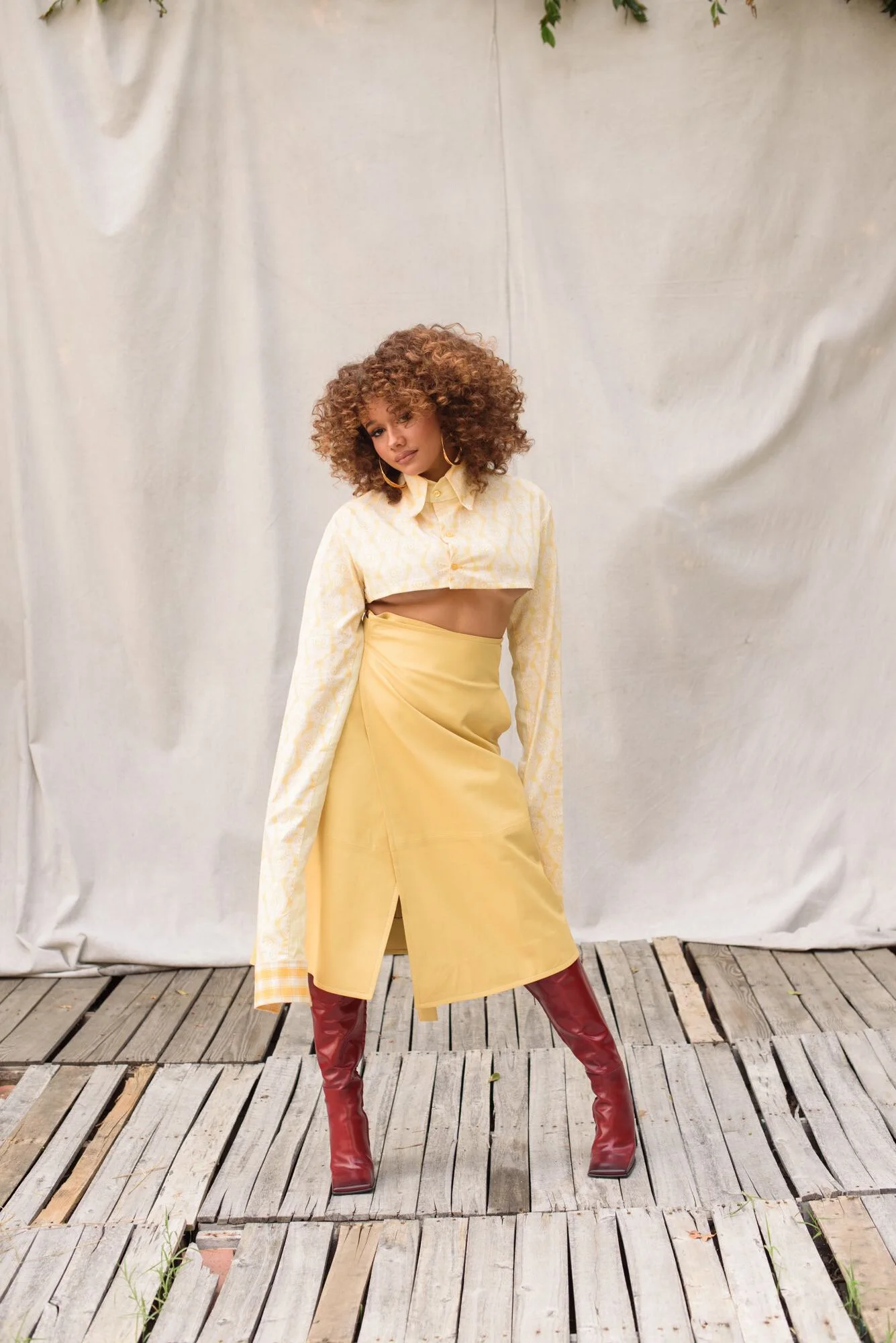 Person with curly hair in yellow cropped top and skirt, standing on wooden platform with a light background, wearing red boots.