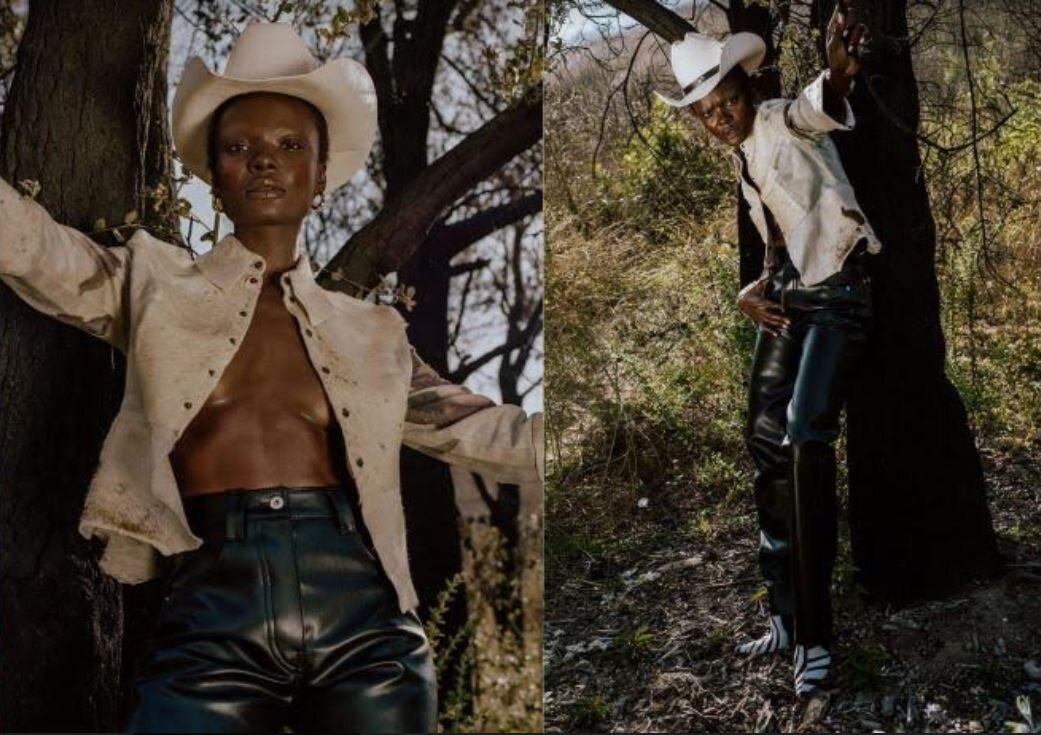 Person wearing a white cowboy hat, open beige jacket, and black leather pants standing outdoors near trees.