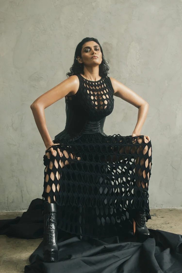 Person wearing a black dress with circular cutouts and black boots, posing against a textured wall.