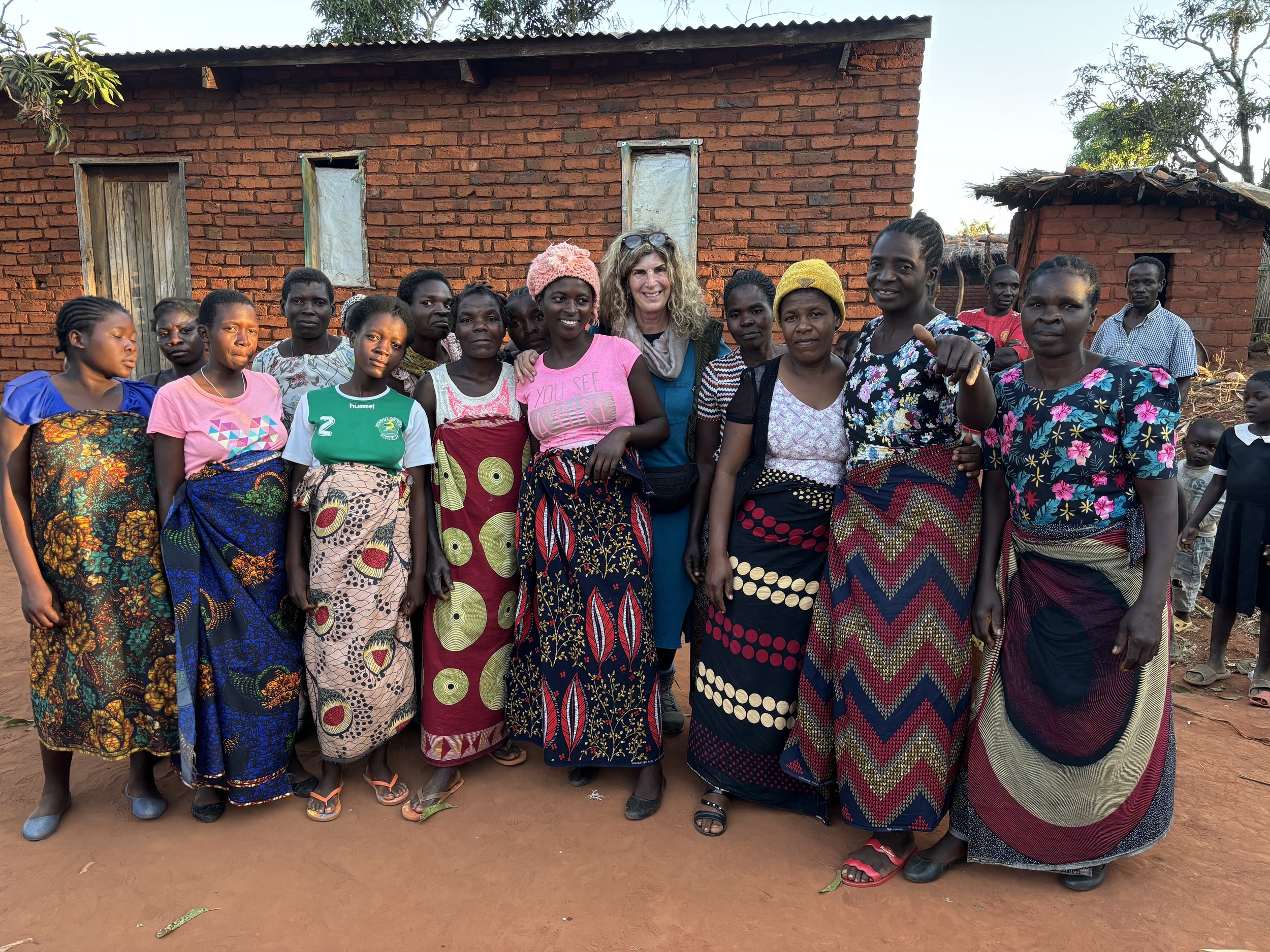 A group of women and children standing outside in front of a brick building, dressed in colorful clothing, with sunny weather and trees in the background.