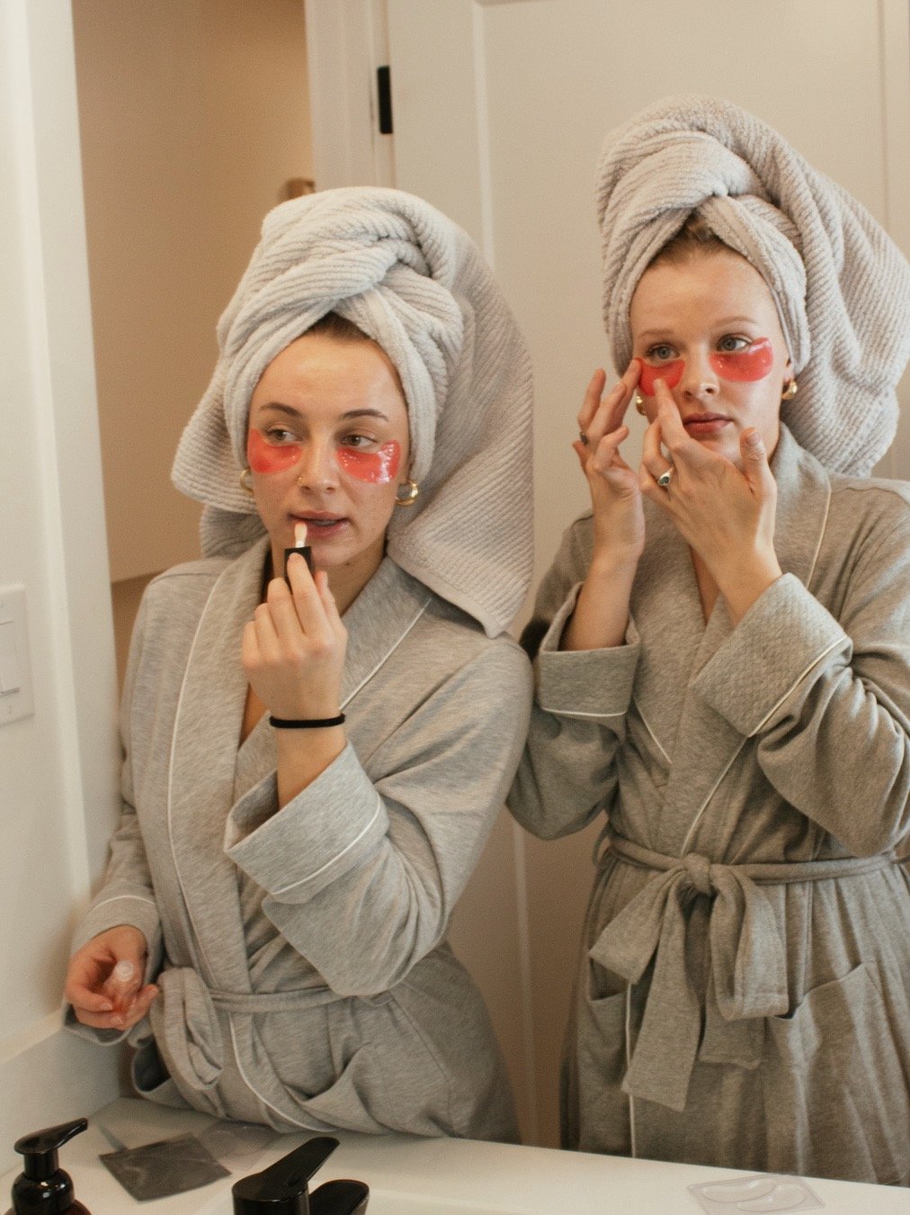 Two women standing in front of a mirror, applying skincare masks on their faces with eye patches underneath. They are wearing gray robes and have towels wrapped around their heads. The setting appears to be a bathroom with personal care items on the counter.