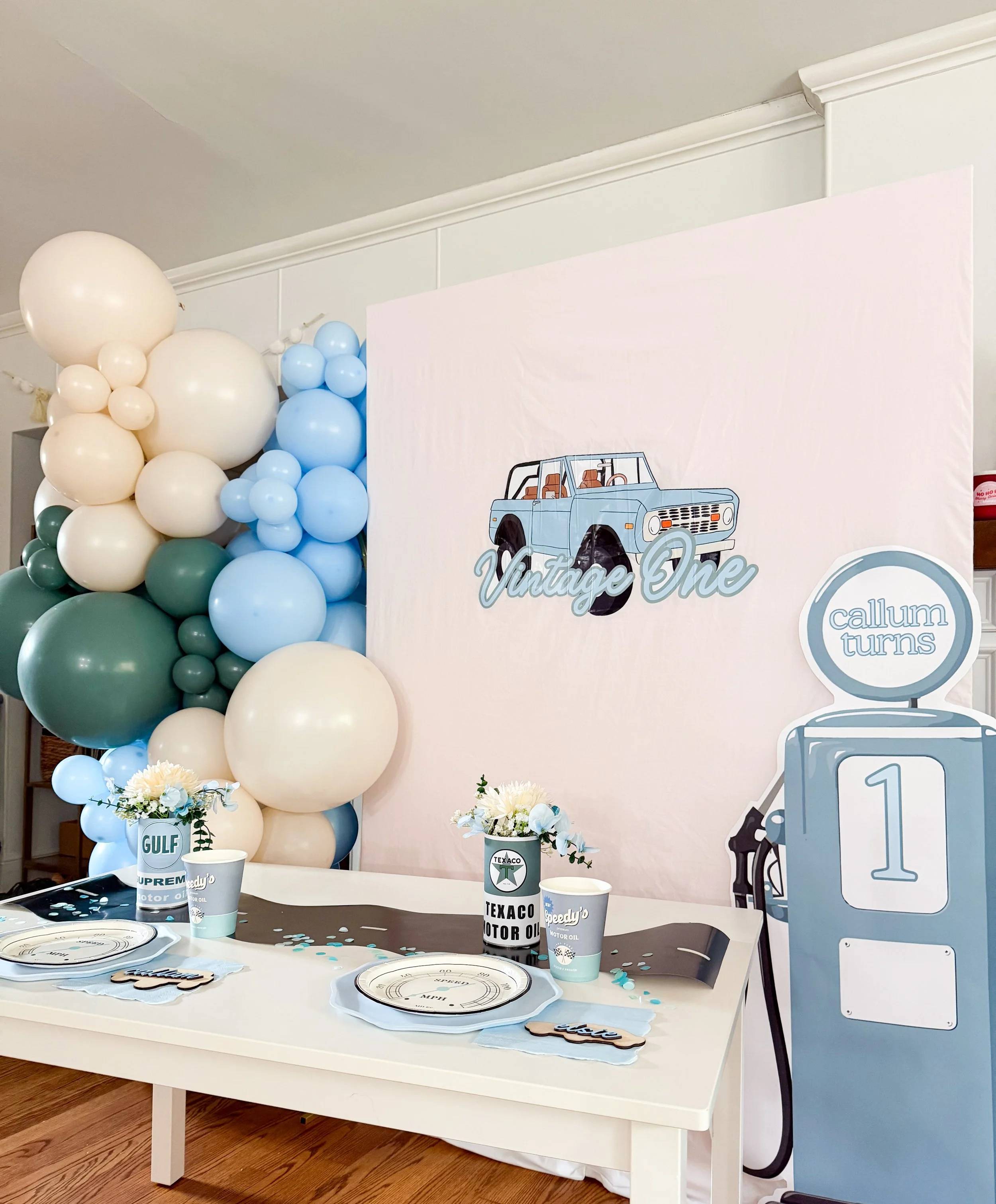 Vintage car-themed first birthday backdrop featuring a classic gas pump, muted blue and green party decor, and nostalgic details.