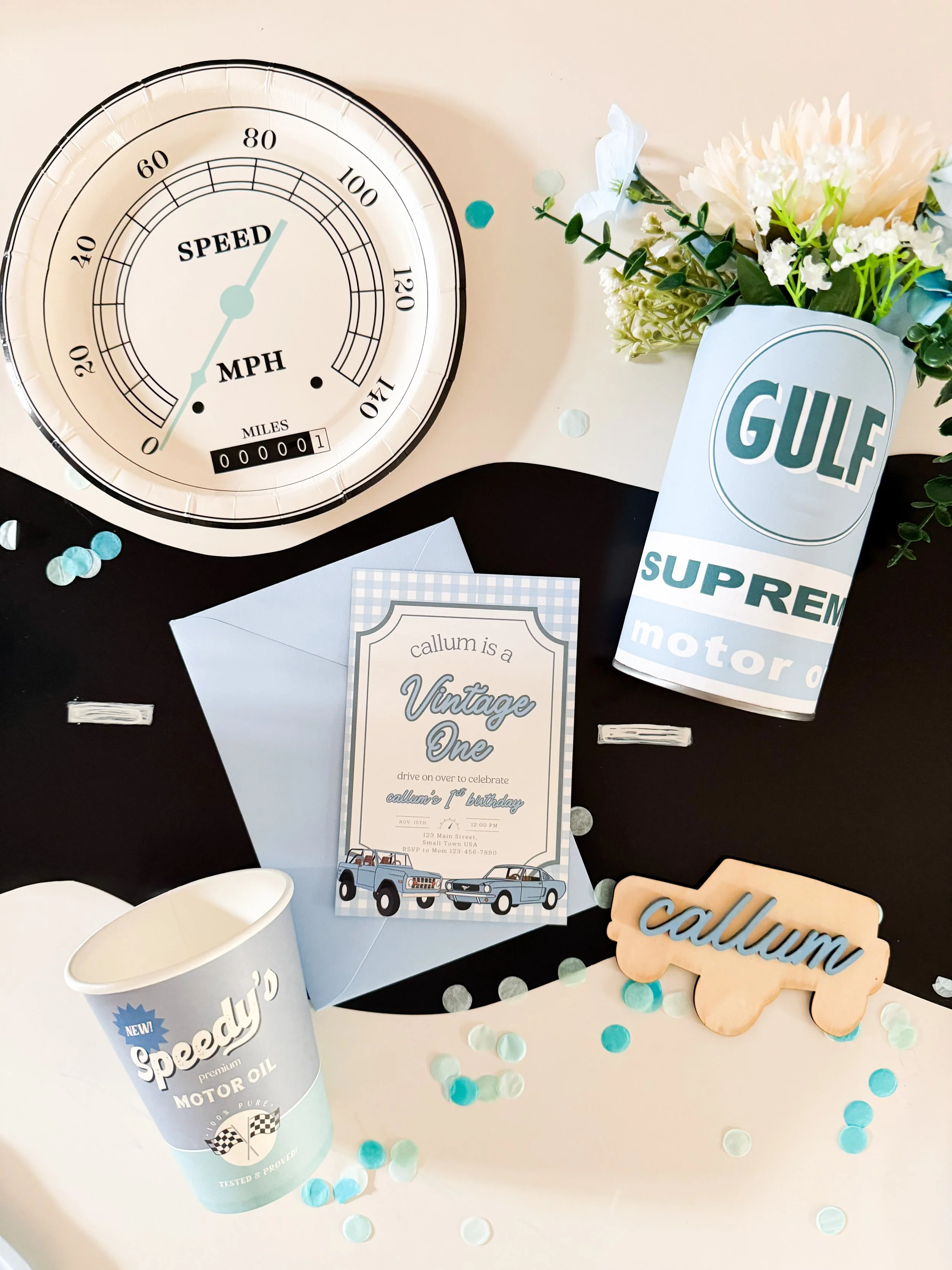 Vintage car-themed first birthday invitation flat lay featuring muted blue and green colors and “Cruising Into One” details.