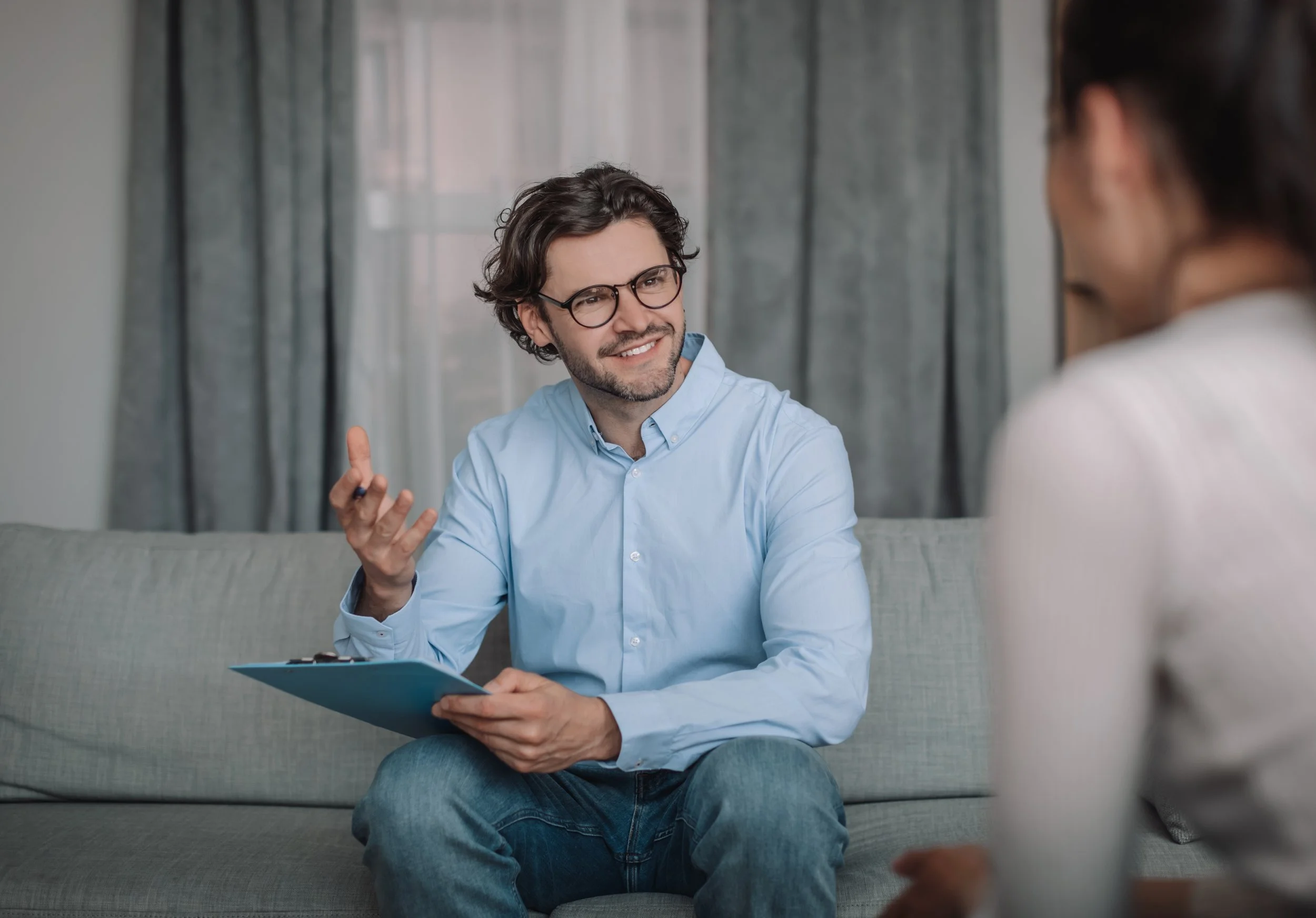 A man with glasses, beard, and curly hair, wearing a light blue shirt, smiling and gesturing with his right hand while holding a clipboard, talking to a woman whose back is turned to the camera, sitting on a couch in a room with curtains.