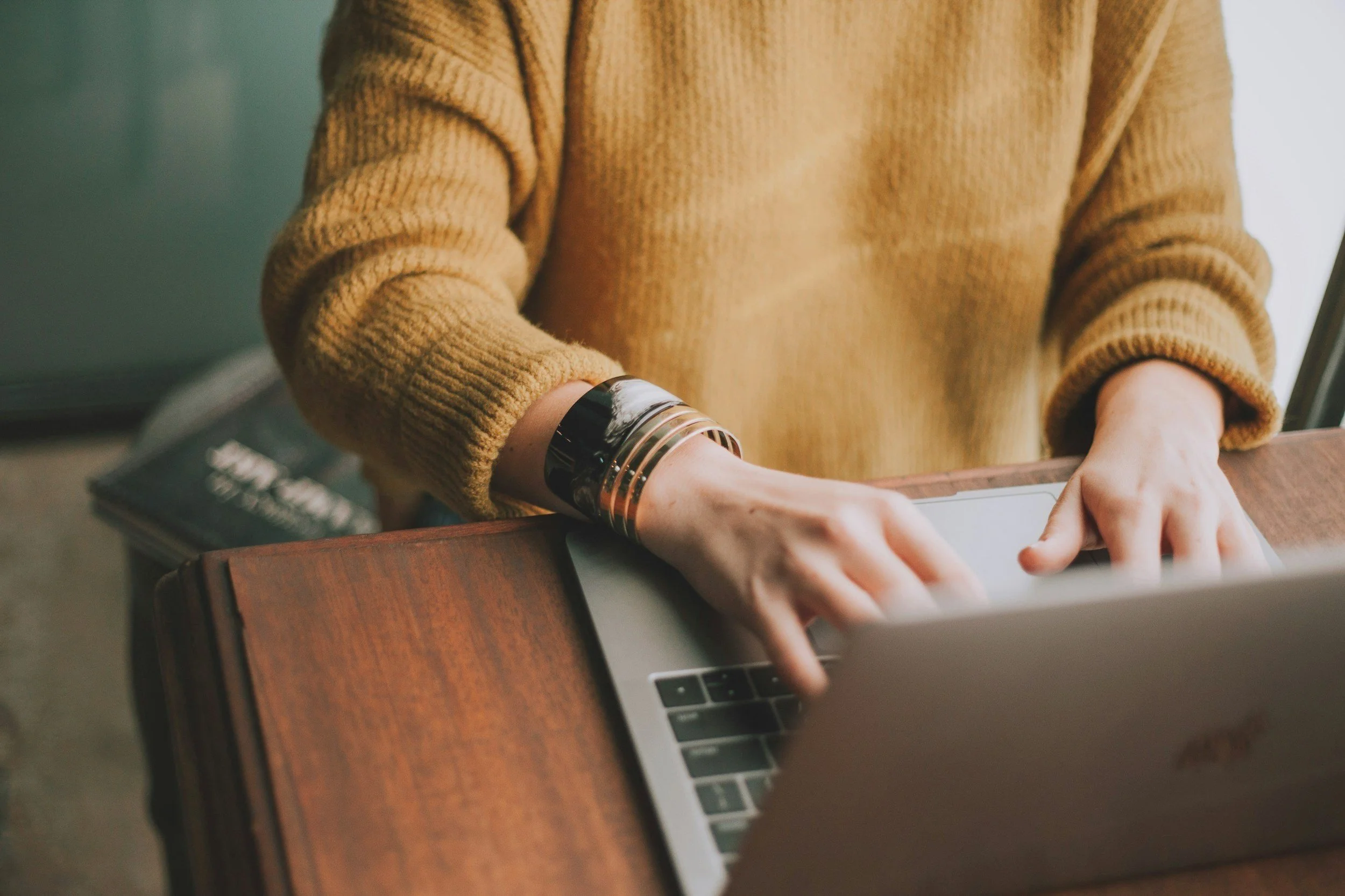 A person wearing a mustard-colored sweater is typing on a silver laptop with their hands resting on the keyboard. They are seated at a wooden table, and a book is visible in the background.