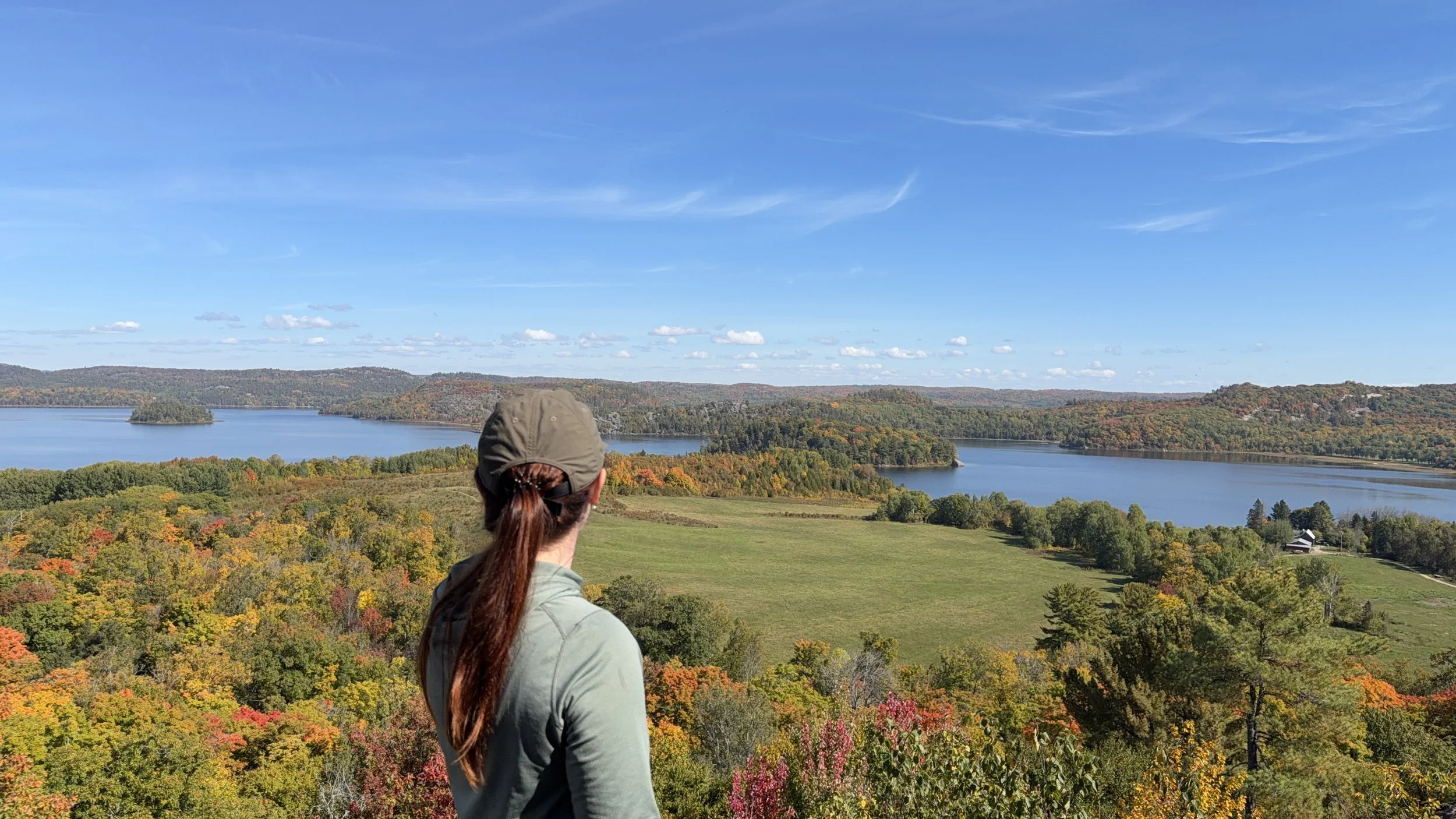 Woman with brown hair and sunglasses taking a photo of a lake and forested hills with her smartphone from a scenic overlook.