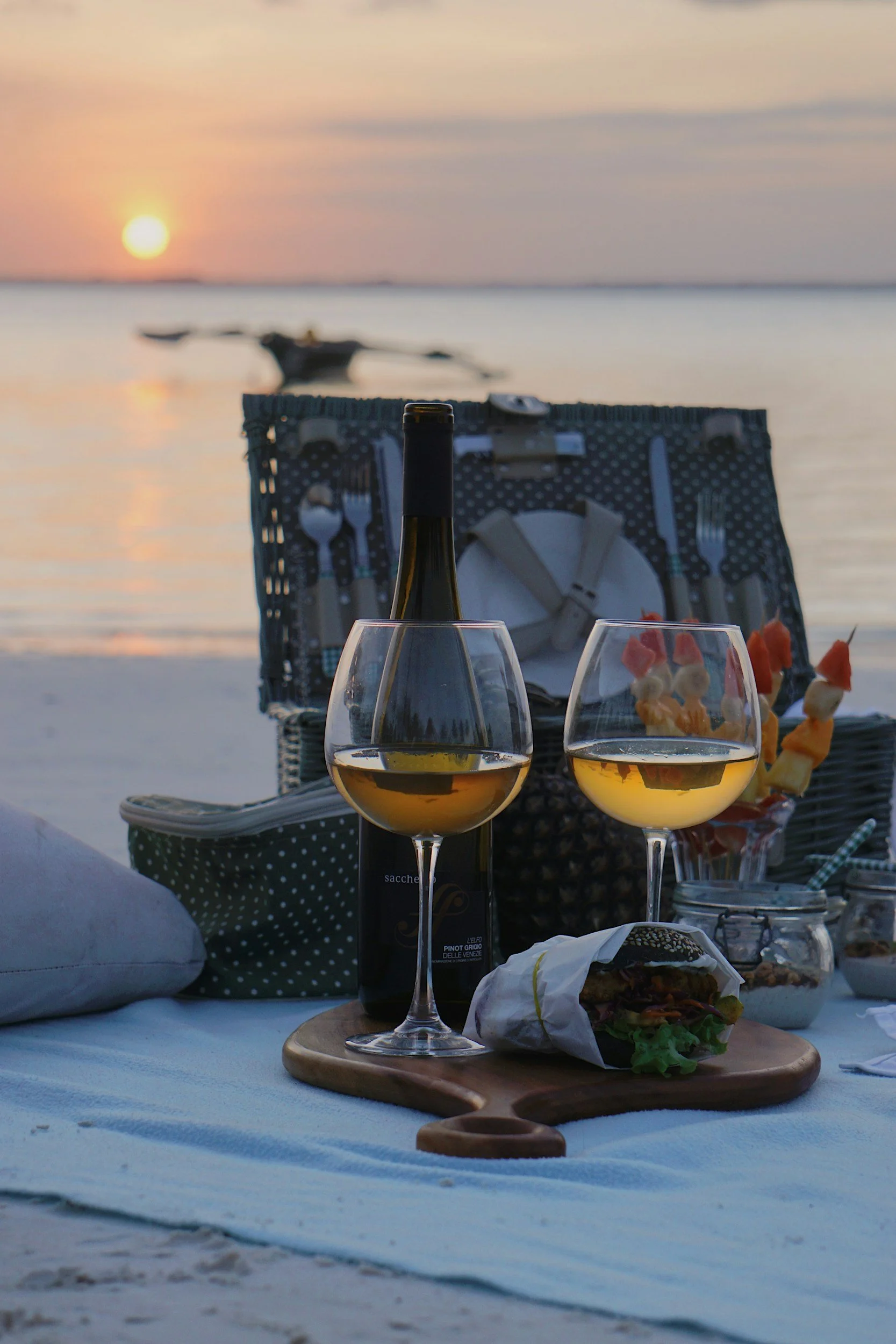 Beach picnic setup at sunset with wine, glasses, food, and basket.