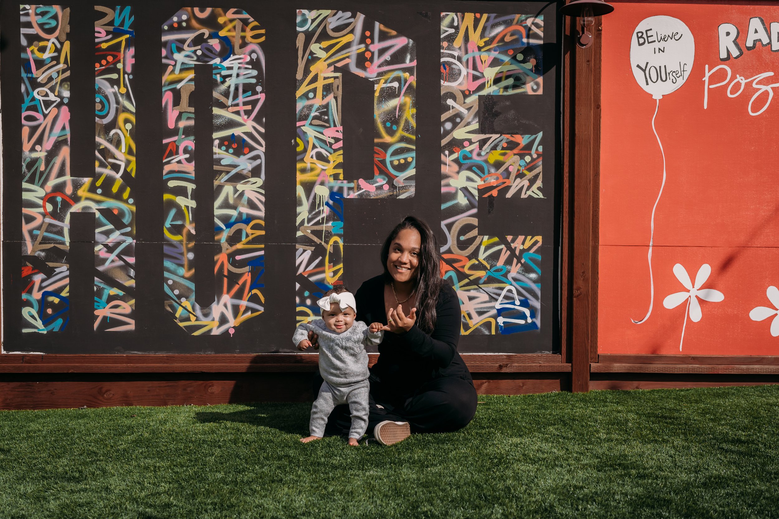 Woman sitting on grass holding a baby in front of a large colorful mural spelling 'HOPE' with graffiti art. Beside the mural, there is a red section with a balloon illustration and the text 'Believe in Yourself.'