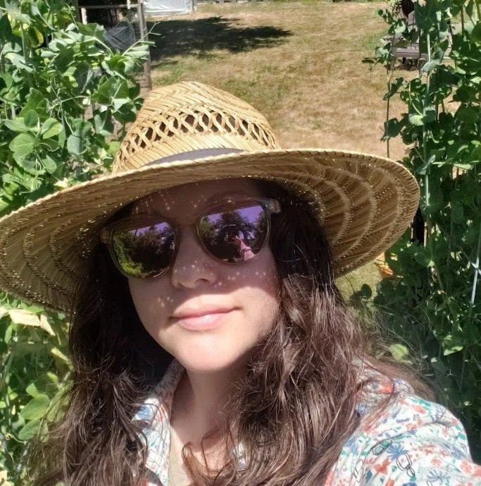 Woman wearing a straw sun hat and large sunglasses standing outdoors among green bushes, with sunlight and shadows on her face.
