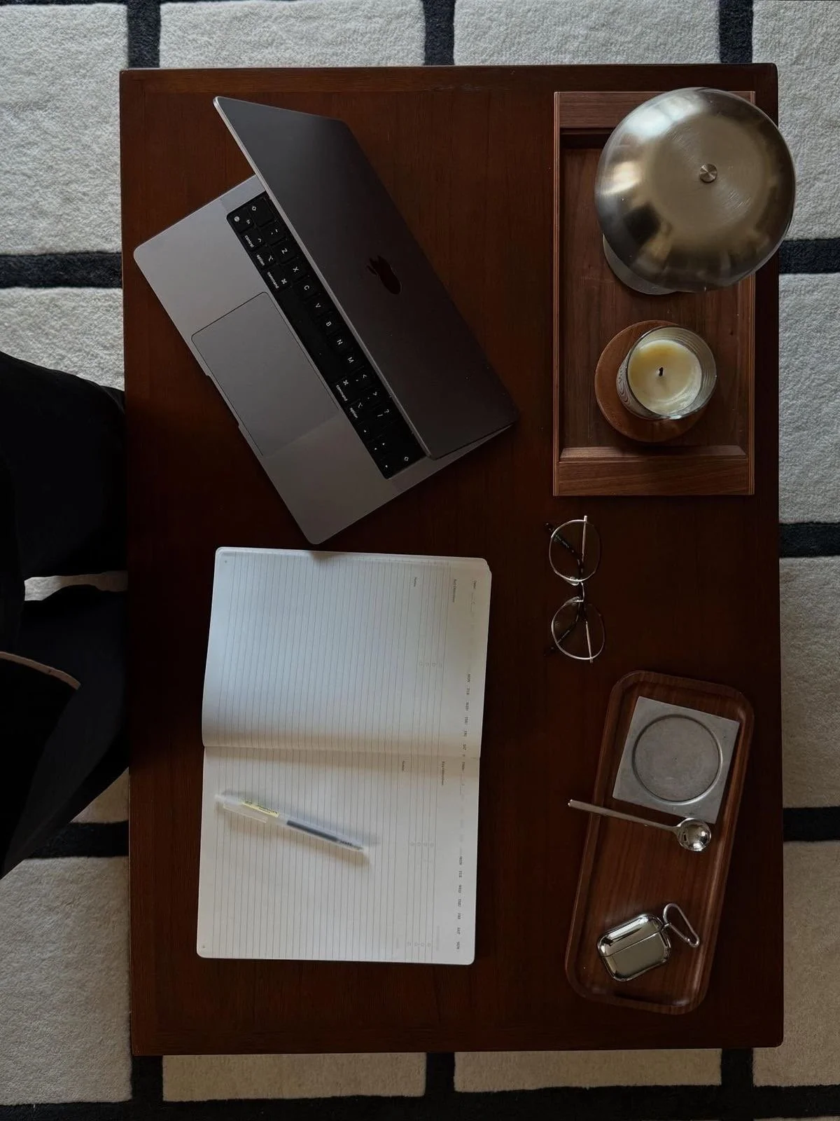 A wooden desk with a silver laptop, a pair of glasses, an open notebook with a pen, a wooden tray holding a silver bowl, a candle, and a small salt shaker, and a gray and cream striped rug underneath.