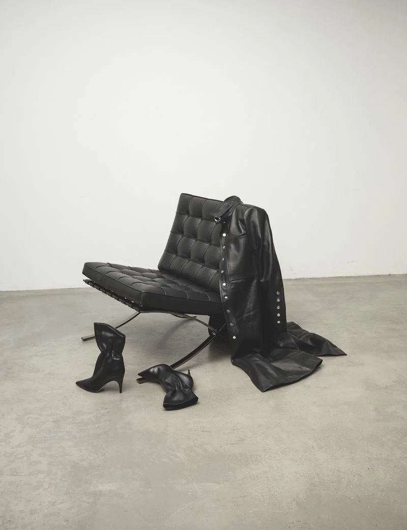 An art installation featuring a black leather chair with a jacket draped over it, a pair of black high-heeled shoes, and a pair of black leather boots, all arranged on a plain concrete floor against a white wall.