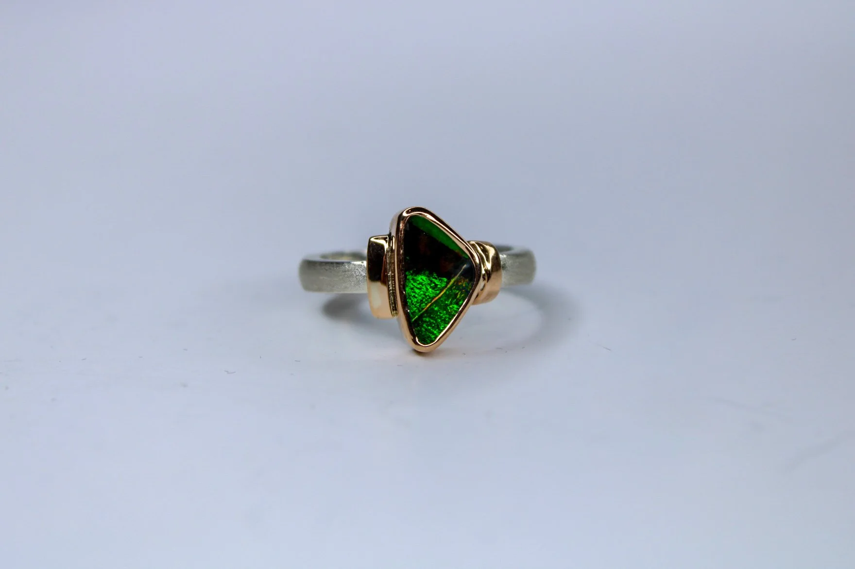 A silver ring with a green and black triangular boulder opal set in a gold bezel on a plain white background.