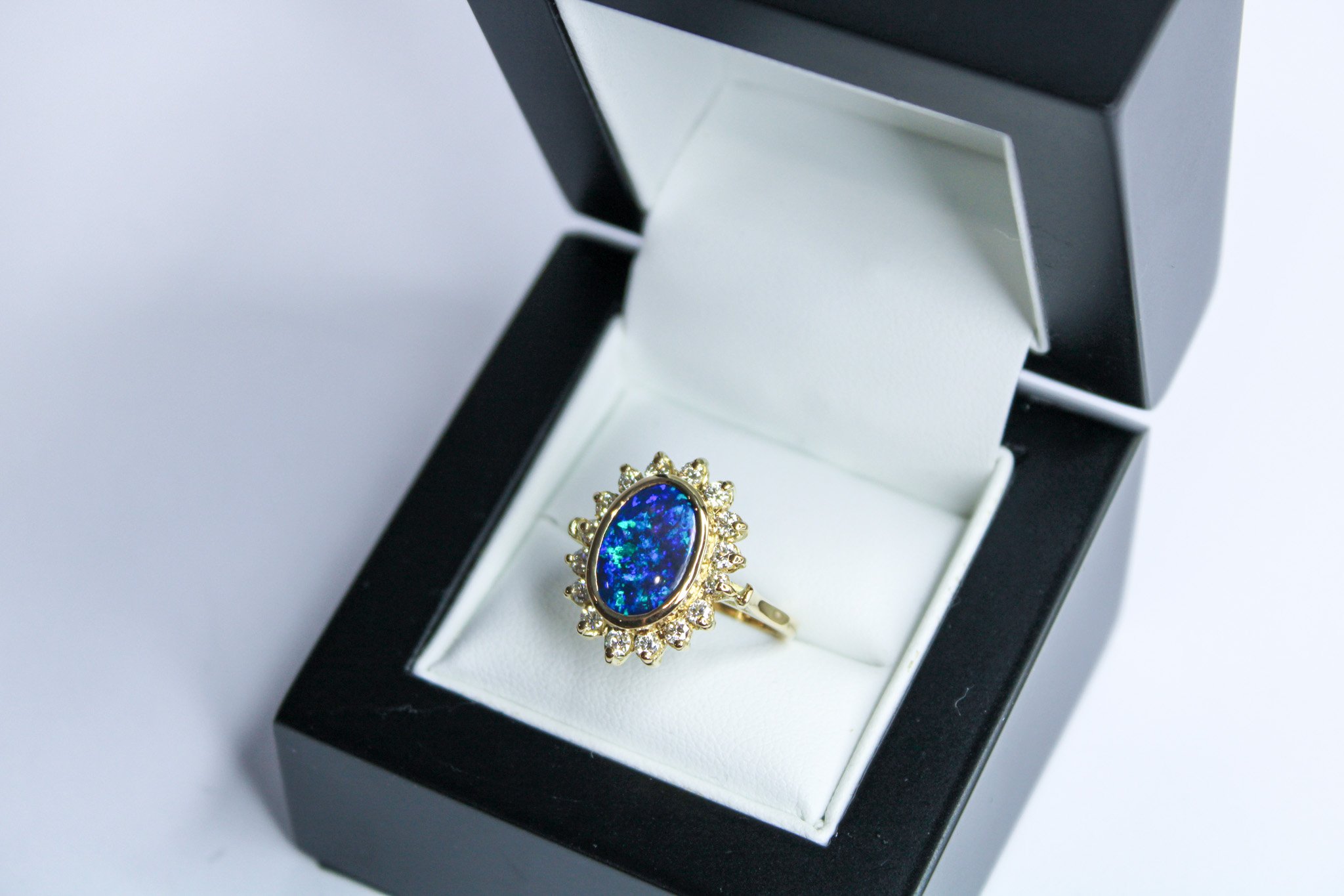 Gold ring with an oval blue opal gemstone surrounded by small diamonds, inside a black jewelry box with white interior.