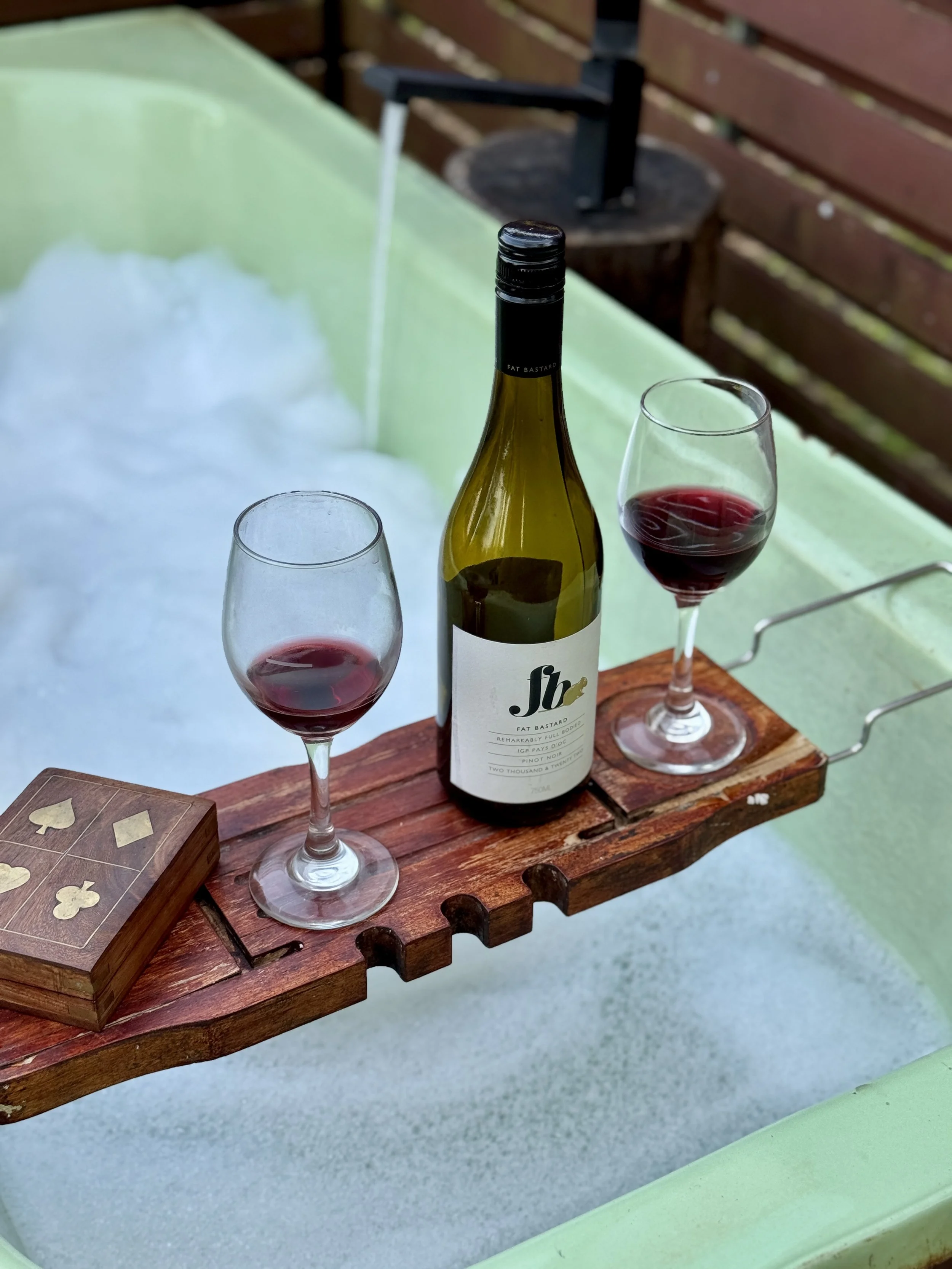Two glasses and a bottle of red wine rest on a plank over a bath filled with bubbles.