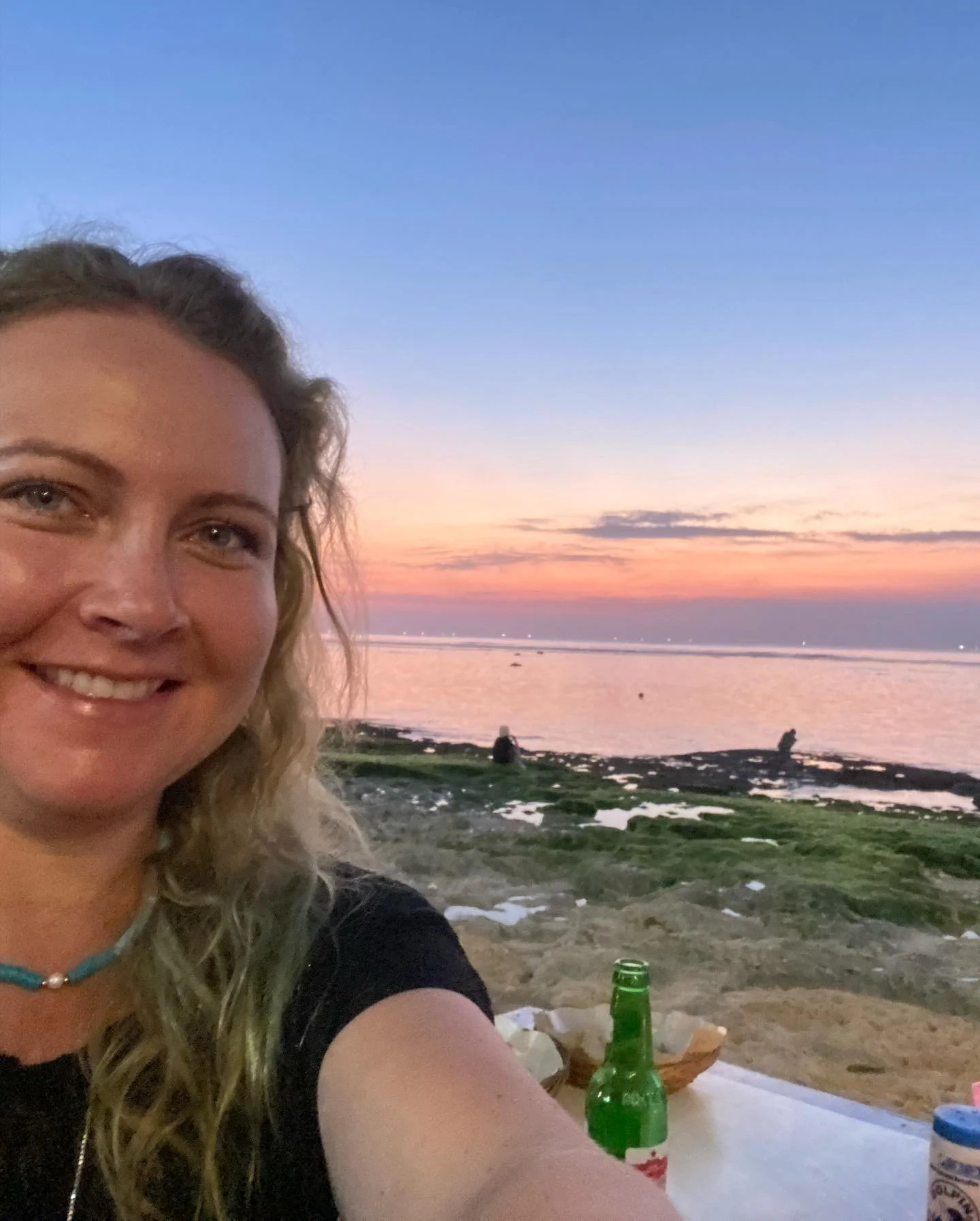 A woman smiling takes a selfie at the beach during sunset with a colorful sky, rocks, and people in the background, and a green beer bottle on the table in the foreground.