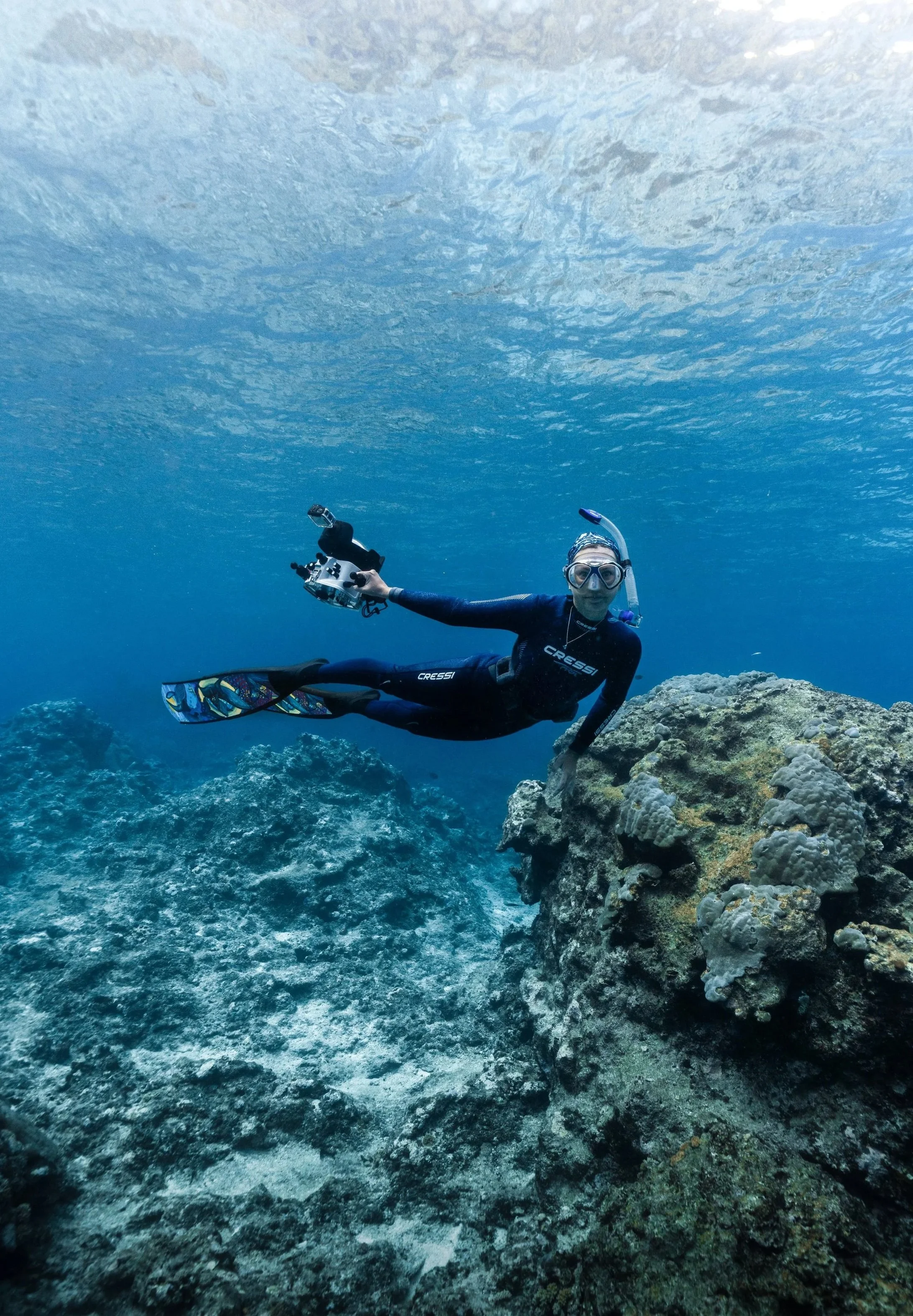 A scuba diver in a black wetsuit, mask, and fins swimming underwater near coral and rocks.