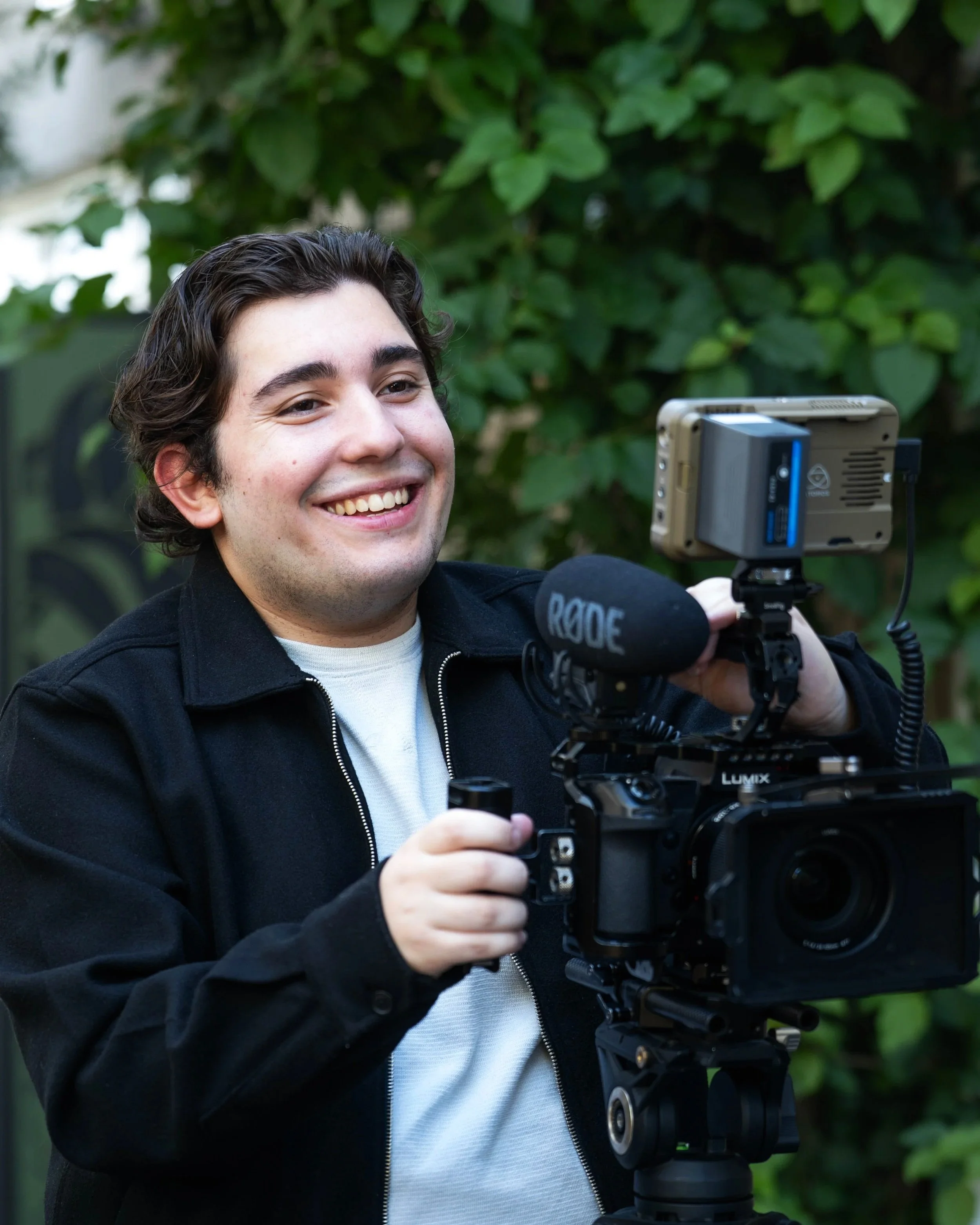Young man smiling while adjusting a professional camera mounted on a tripod outdoors with green foliage in the background.