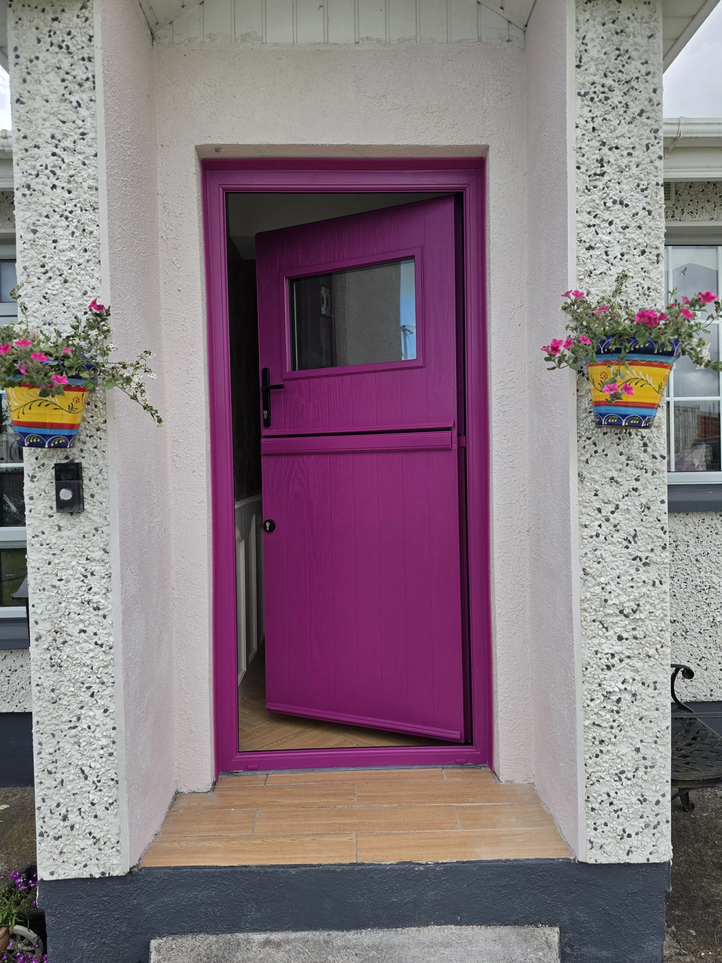 Front door painted bright pink with a small window and black handle, flanked by two hanging flower pots with pink and white flowers on textured exterior walls, with a small sidewalk and stairs in front.