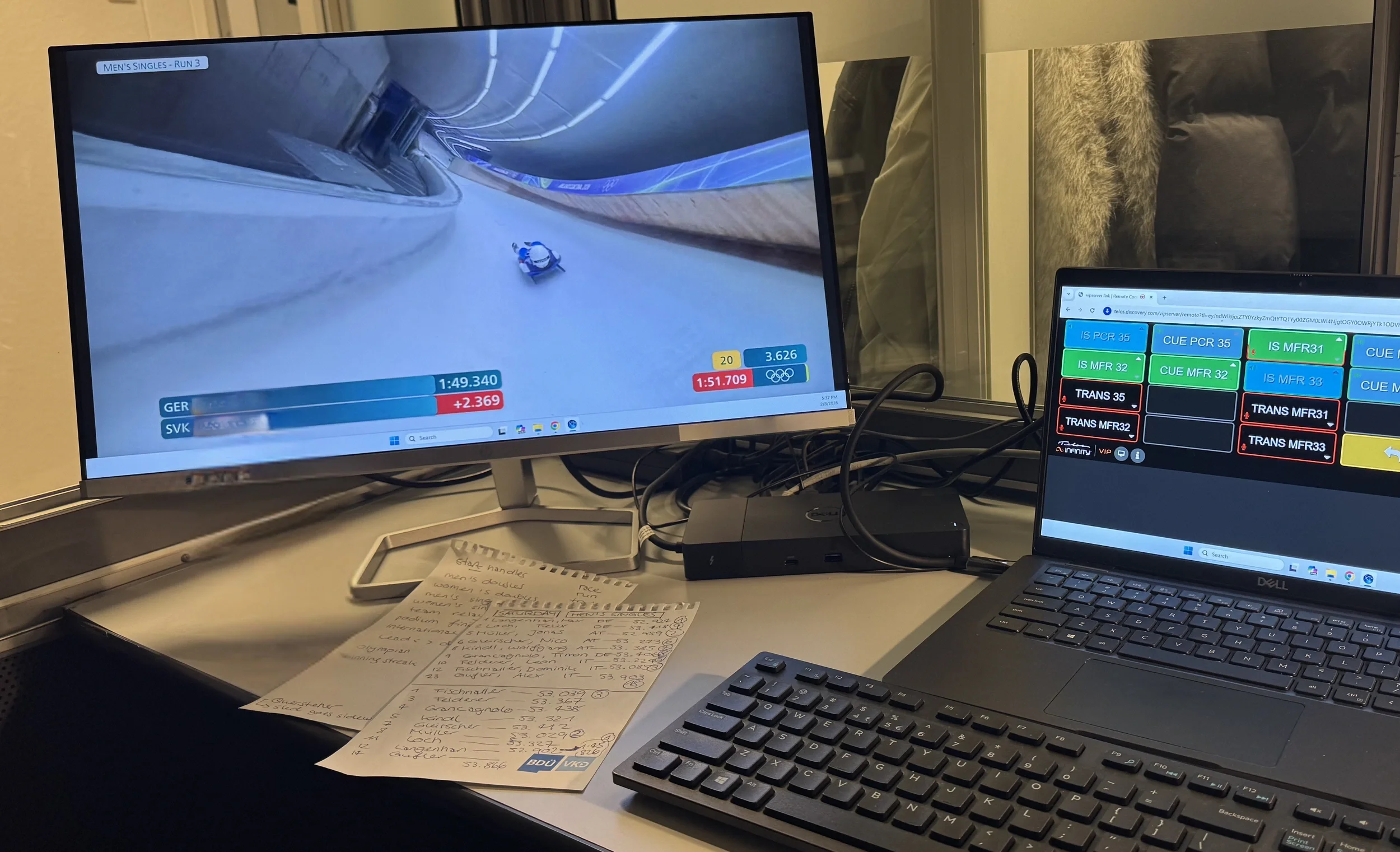 Zwei Bildschirme in einer Dolmetschkabine. Links ist die Live-Übertragung eines Rodel-Wettbewerbs zu sehen, rechts verschiedene Übertragungskanäle. Auf dem Tisch liegt ein Notizblock mit Vokabellisten.