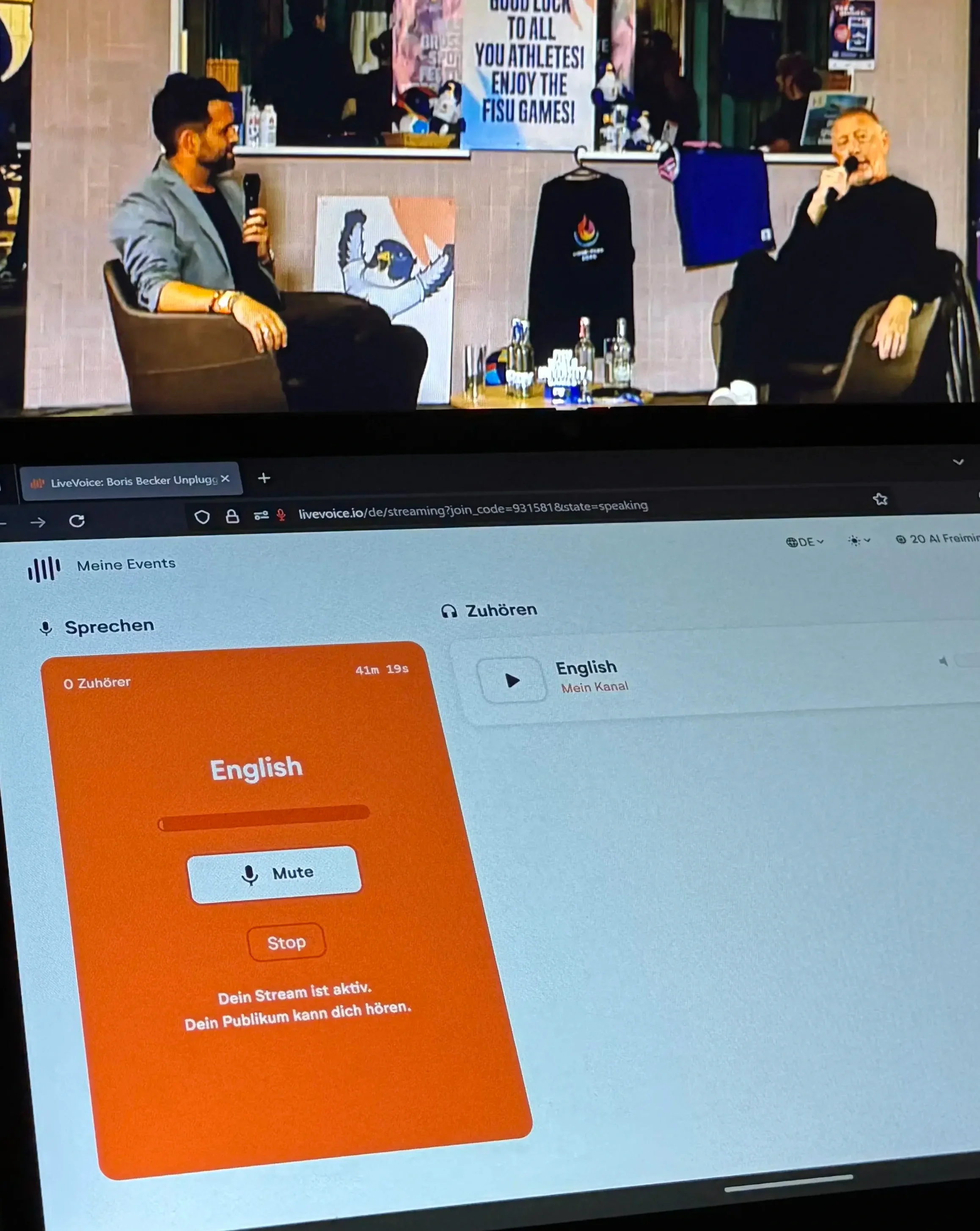 Auf einem Podium spricht ein Moderator mit Boris Becker. Im Vordergrund ist ein Bildschirm zu sehen, auf dem eine Dolmetschplattform läuft. Unter einem Kopfhörersymbol steht: Zuhören, Englisch, Mein Kanal.