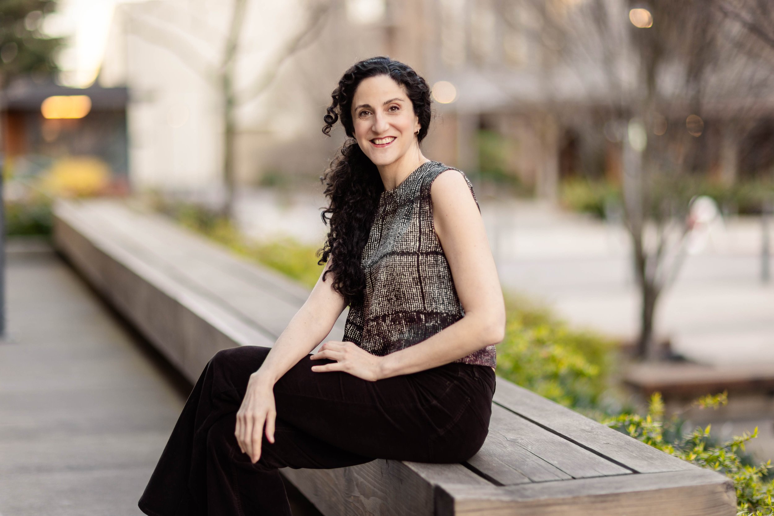 A woman with curly black hair and a big smile, standing outdoors next to a black brick wall, wearing an olive green t-shirt, beige pants, and a smartwatch.