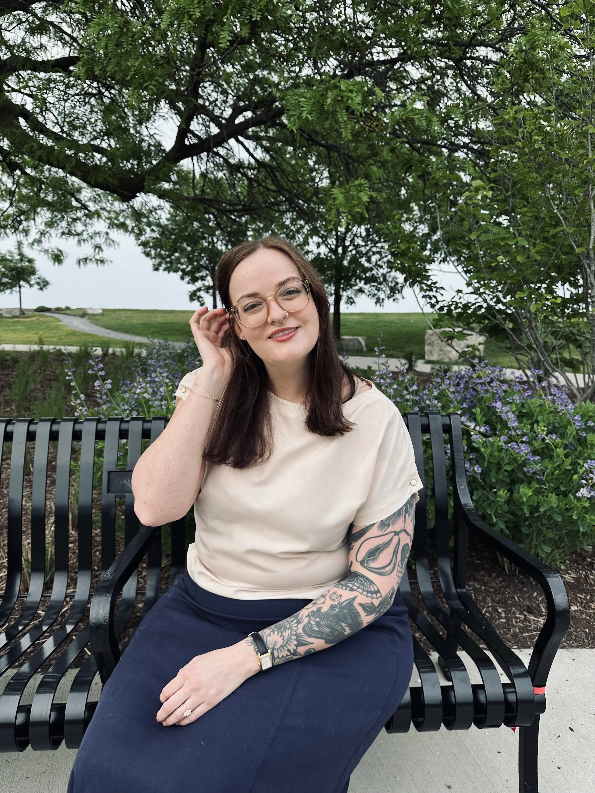 Person sitting on a park bench with green trees and purple flowers in the background.