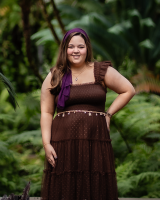 A woman standing outdoors in a lush green garden, smiling, wearing a brown dress with purple accents and a purple scarf.