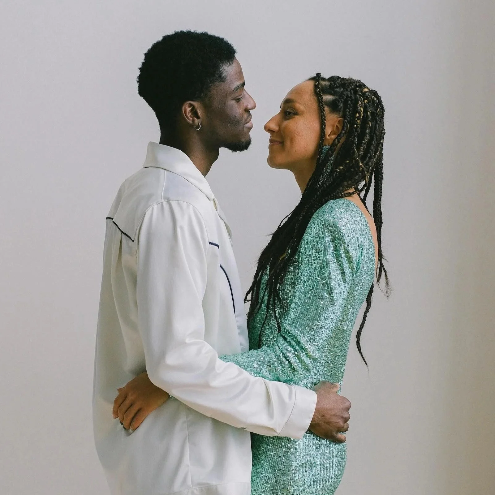 A man and woman face each other closely, smiling and holding hands, in front of a plain white wall.