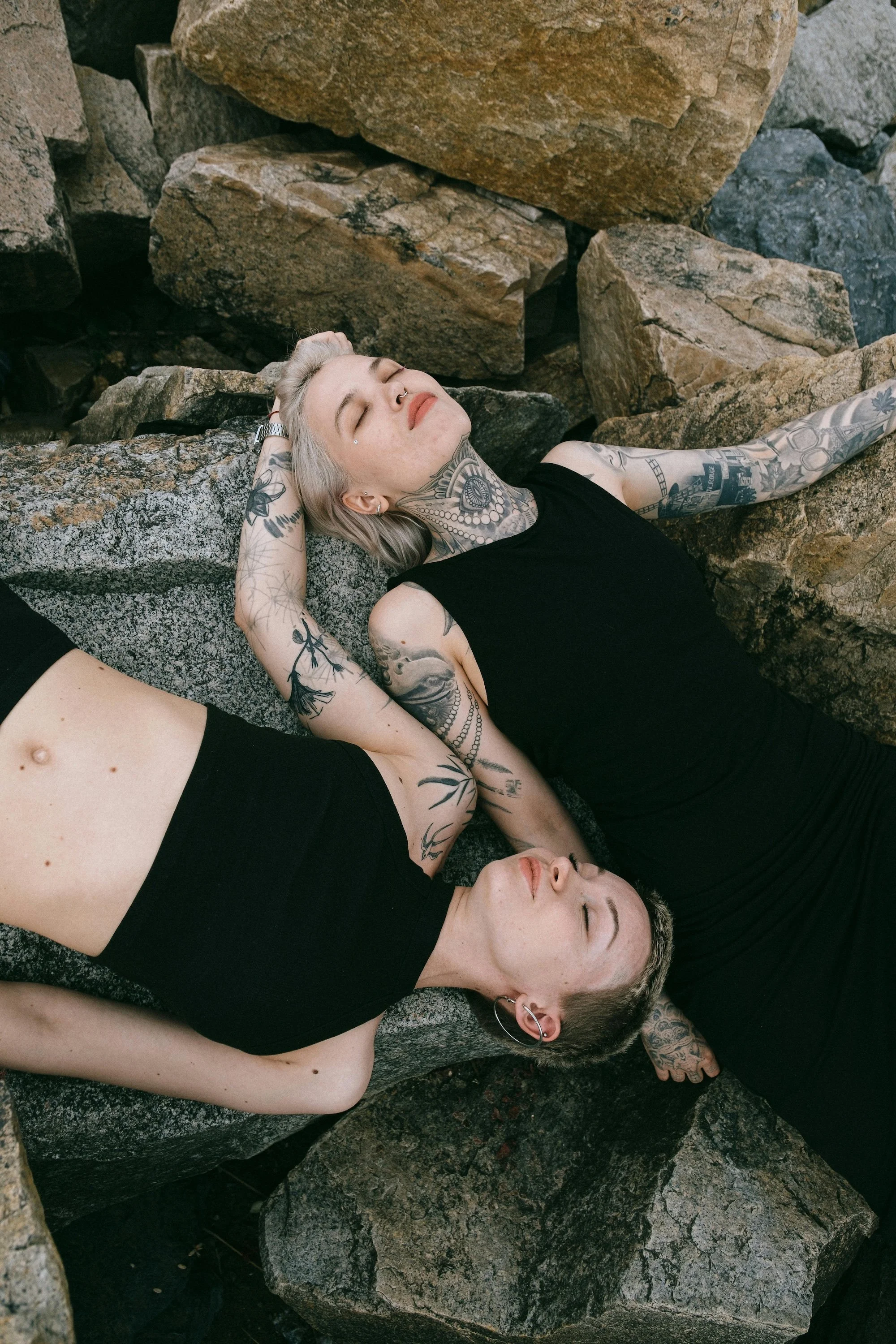 Two young women with tattoos are lying down on large rocks with their eyes closed, appearing relaxed and peaceful.