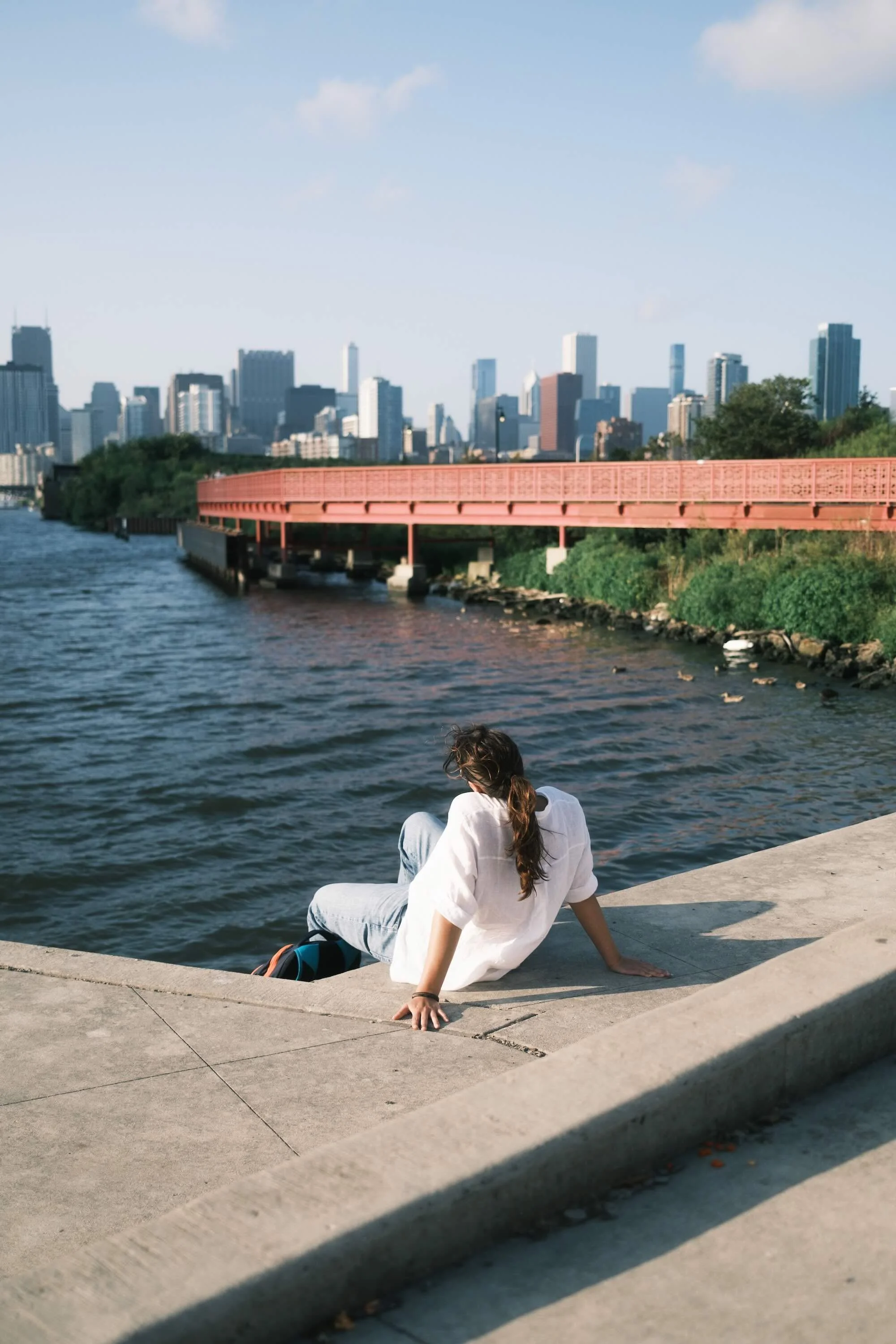 A woman with long brown hair, wearing a white shirt and light blue jeans, sitting on a concrete ledge by the water with her back to the camera, overlooking the Chicago skyline.