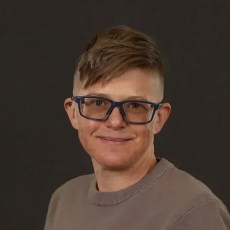 Portrait of a young man with light brown hair, glasses, and a friendly expression, wearing a gray shirt against a dark background.