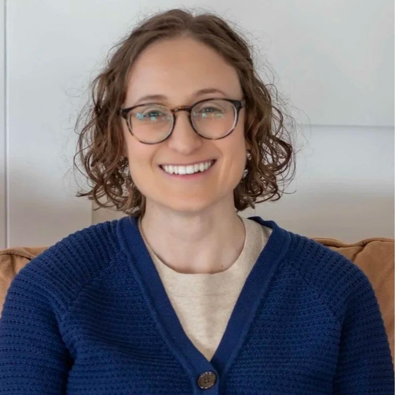 A woman with curly brown hair, glasses, and a friendly smile, wearing a blue sweater and a beige shirt, sitting in a room with a white wall background.