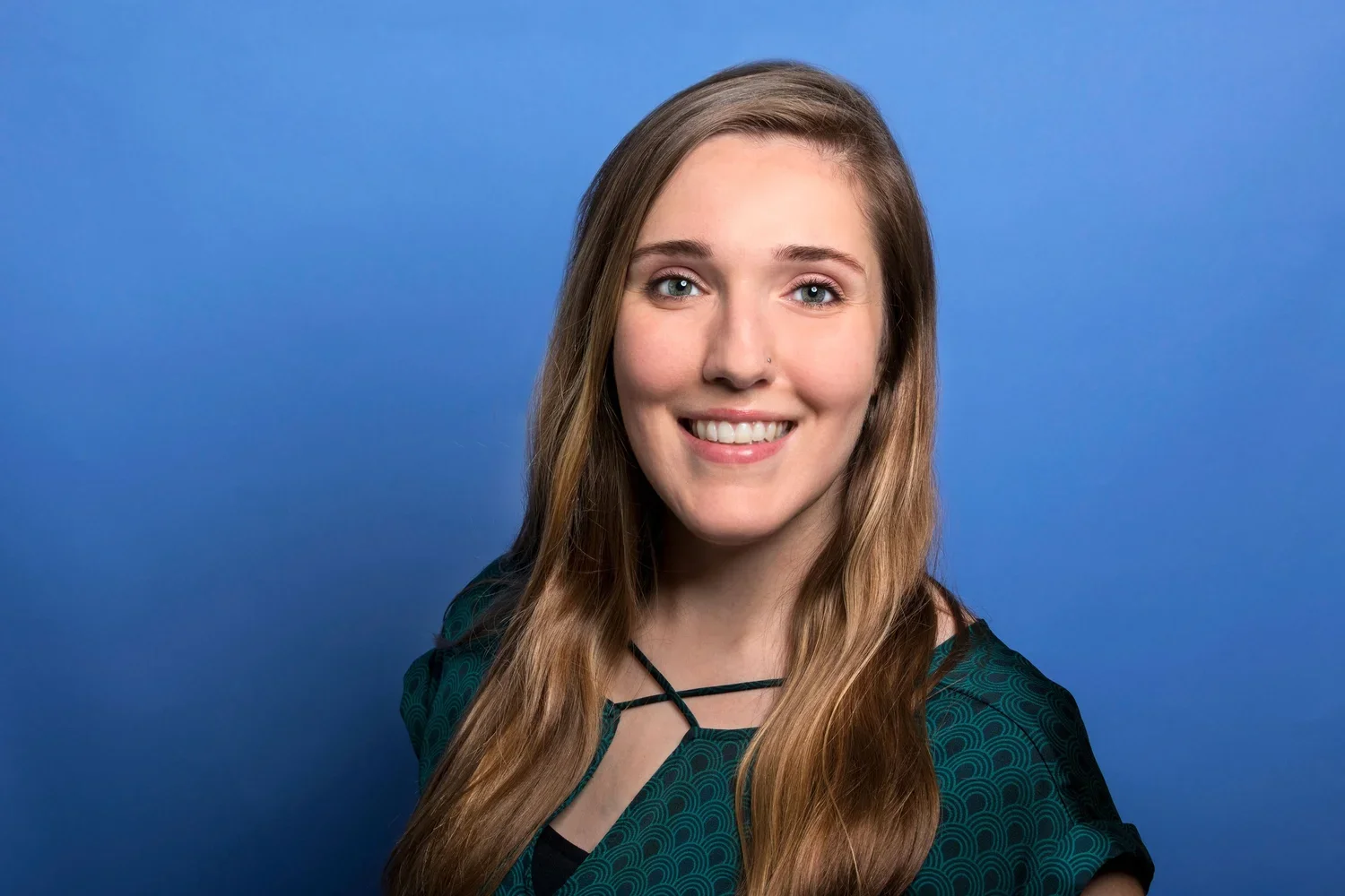 A young woman with long, wavy brown hair, bright blue eyes, and a friendly smile, standing against a plain blue background.