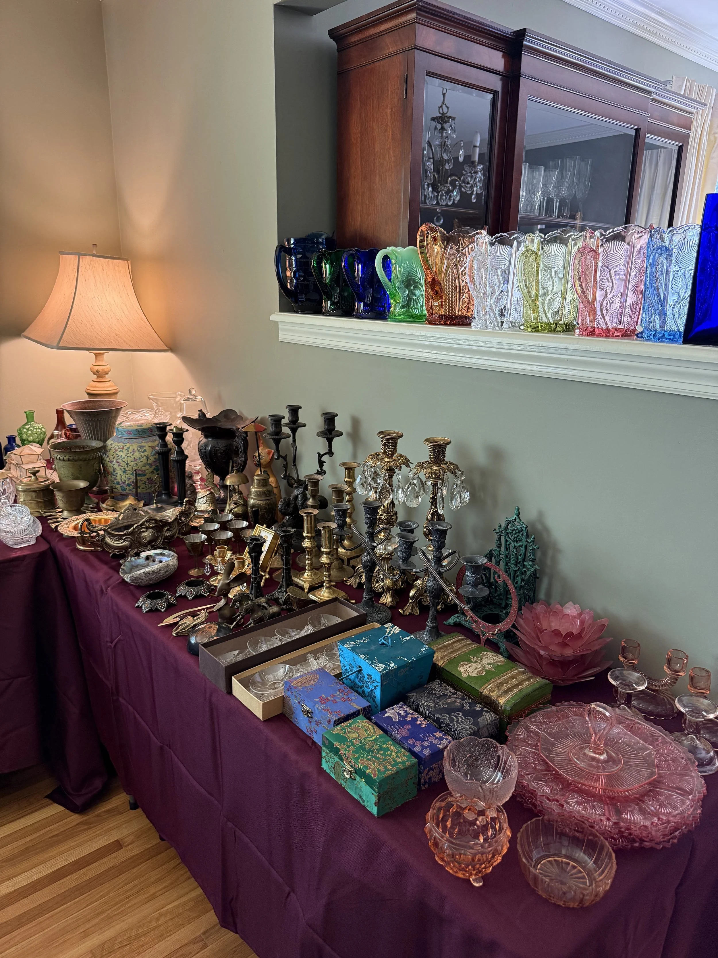 Display of various vintage glassware, candlesticks, jewelry boxes, and decorative items on a purple tablecloth with a wooden cabinet and a lamp in the background.