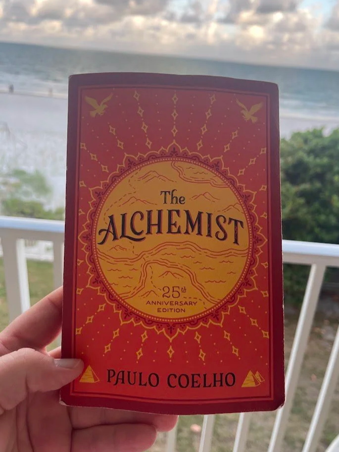 A close-up shot of a hand holding the red and gold 25th Anniversary Edition cover of 'The Alchemist' by Paulo Coelho. The background shows a white balcony railing, green coastal bushes, and a soft view of the ocean.